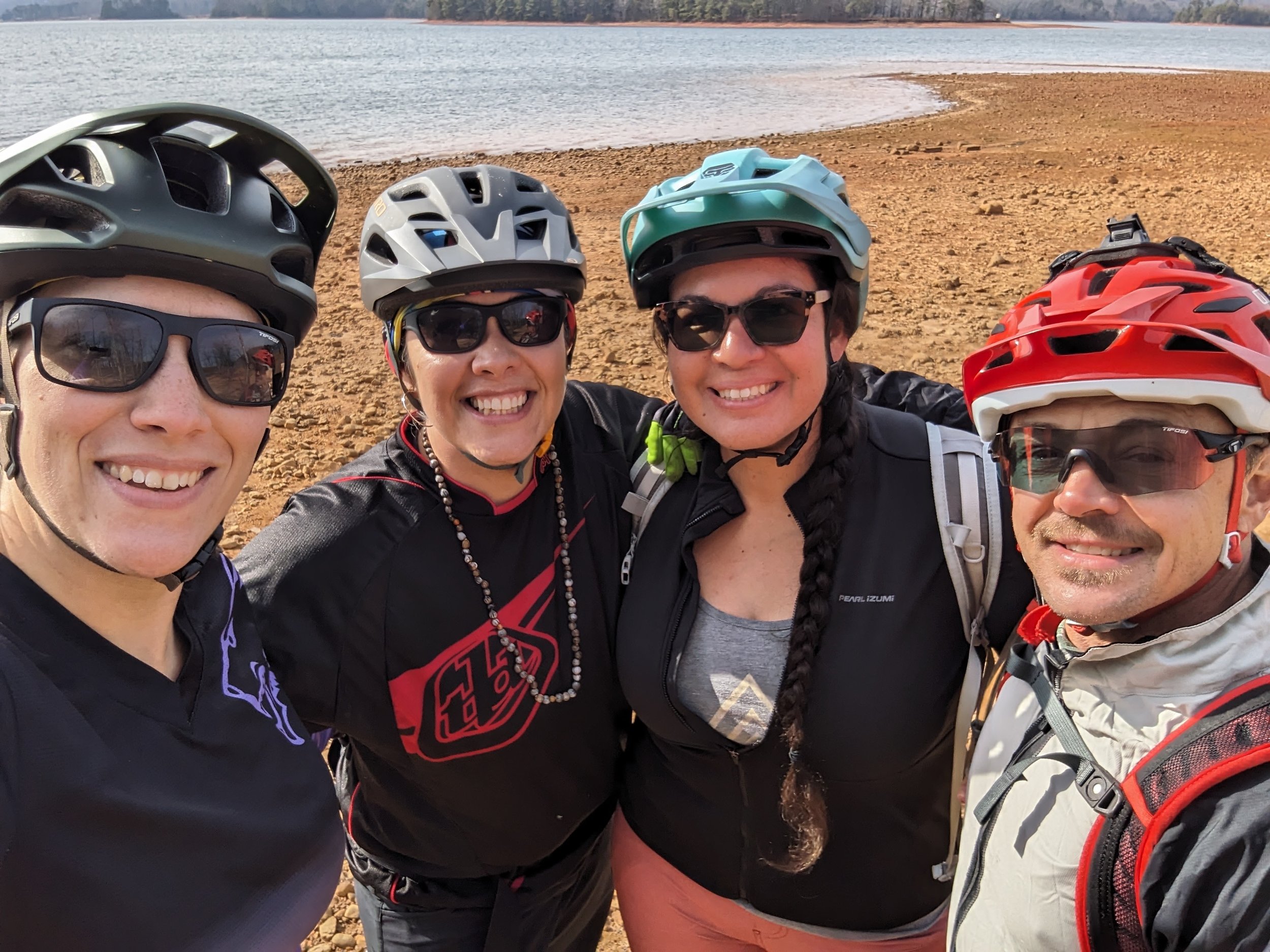Four friends wearing helmets and sunglasses, taking a selfie near a body of water on a sunny day.