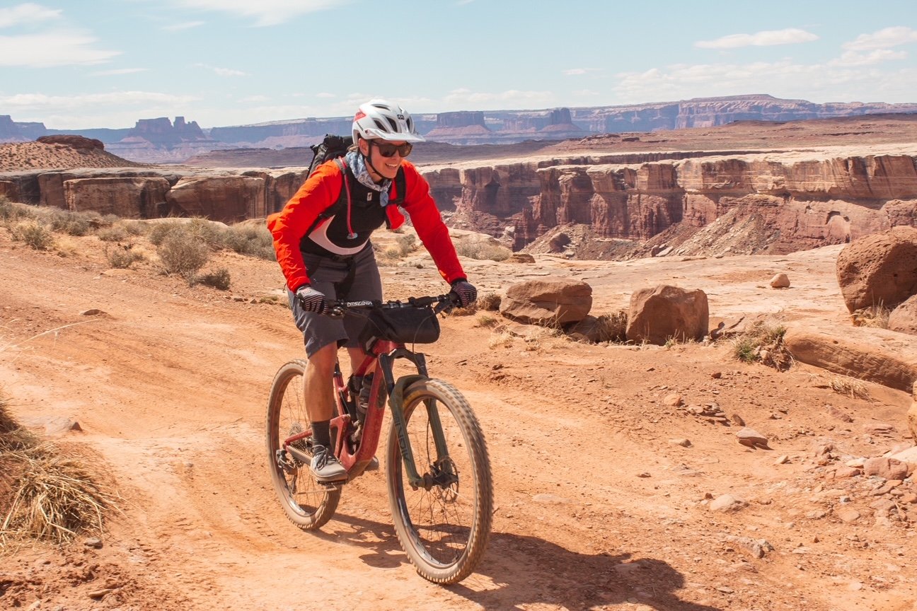 Person wearing a red jacket, helmet, and sunglasses riding a mountain bike on a dirt trail in a desert canyon landscape with large rock formations and blue sky.