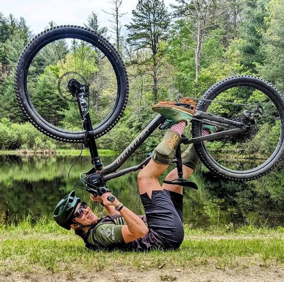 Person lying on the grass by a lake, holding a mountain bike upside down with both wheels in the air, wearing a helmet, sunglasses, and cycling gear, surrounded by lush green trees.