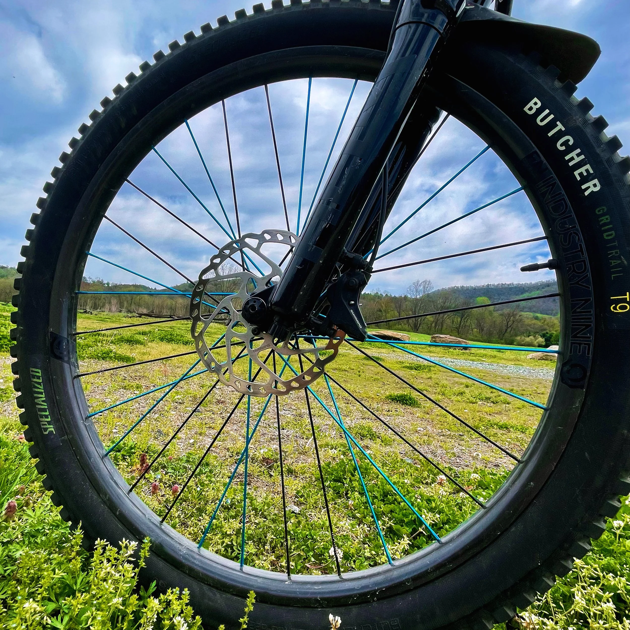 Close-up of a mountain bike wheel with a disc brake and tire, set in a grassy field with a blue sky and distant hills in the background.