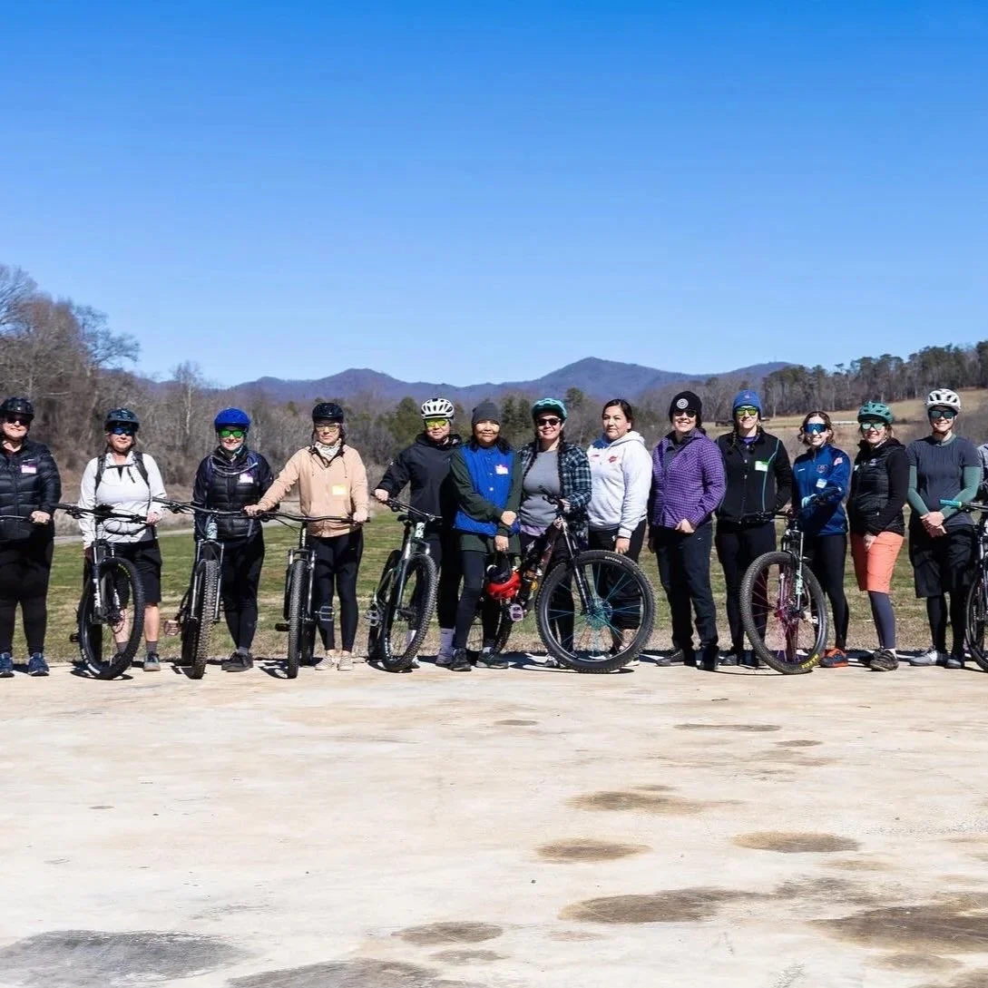 Group of women with bicycles standing outdoors on a sunny day with mountains in the background.