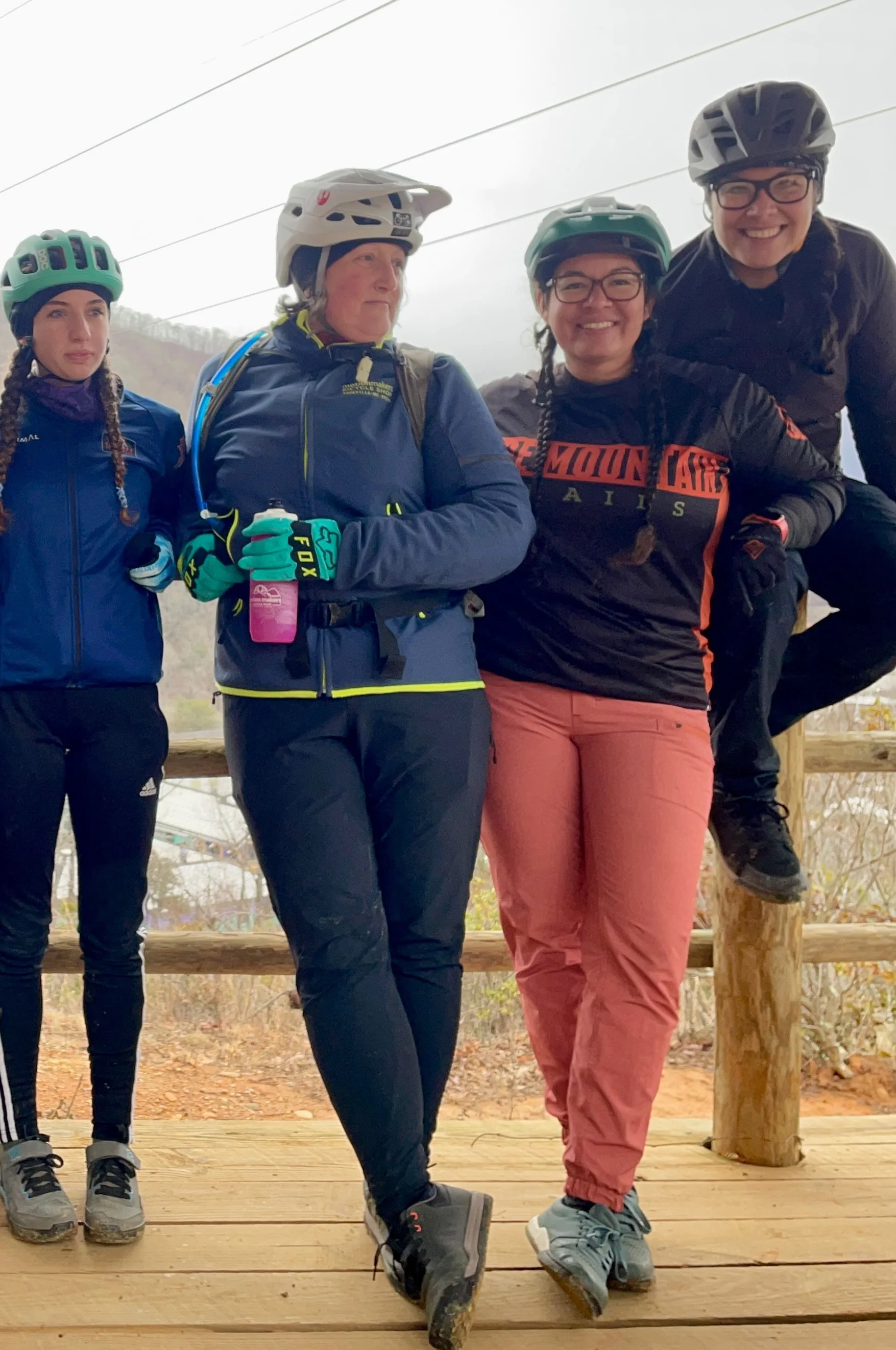 Four women wearing helmets and outdoor gear, standing on a wooden deck with a foggy outdoor background.