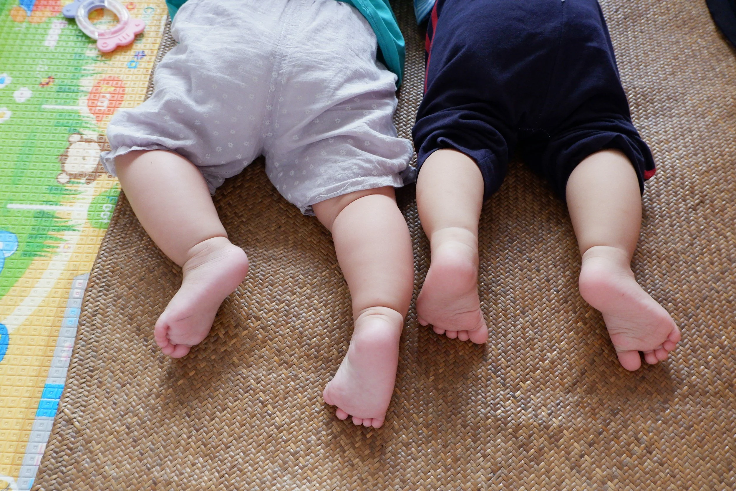Two babies lying on their stomachs on a textured mat with colorful toys nearby, viewed from behind.