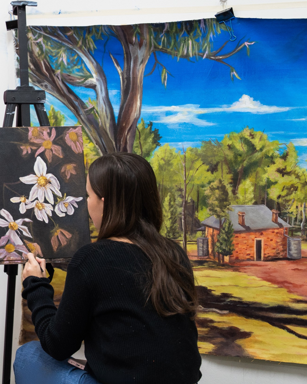 A woman painting a floral scene on a black canvas with a landscape featuring a large tree, a house, and a blue sky in the background.