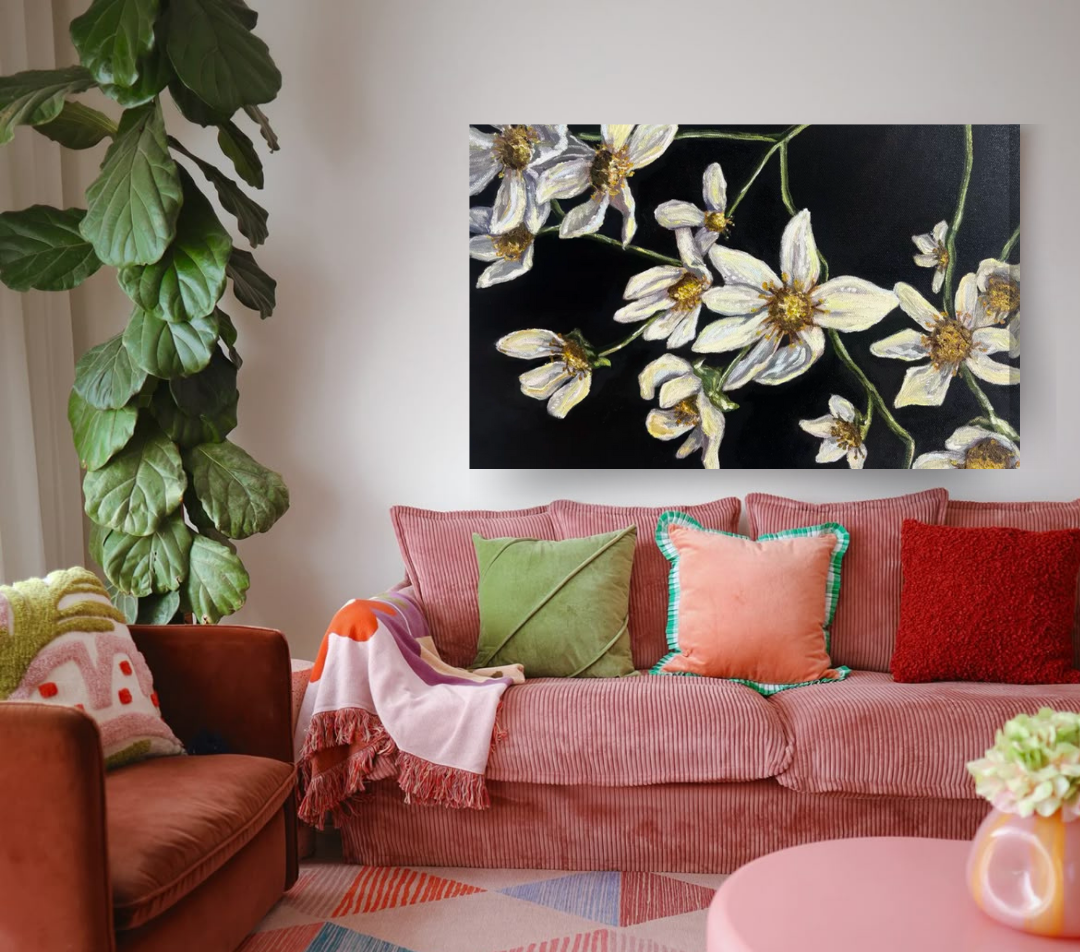 A living room with a pink striped couch decorated with colorful pillows, a large houseplant with broad green leaves on the left, a circular pink table with a flower arrangement in a pale pink vase, a colorful geometric patterned rug, and a painting of white flowers on black background hanging above the couch.