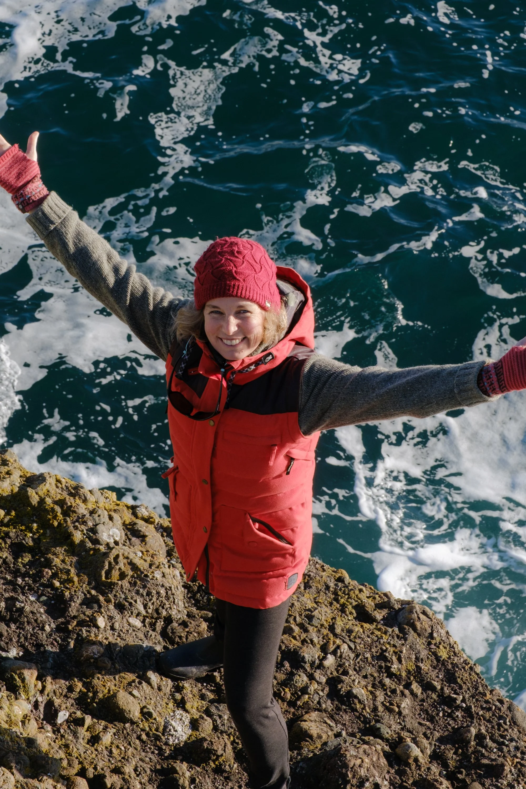 A woman standing on rocky coast with arms outstretched, smiling, wearing a red hat and a red vest, with ocean waves in the background.