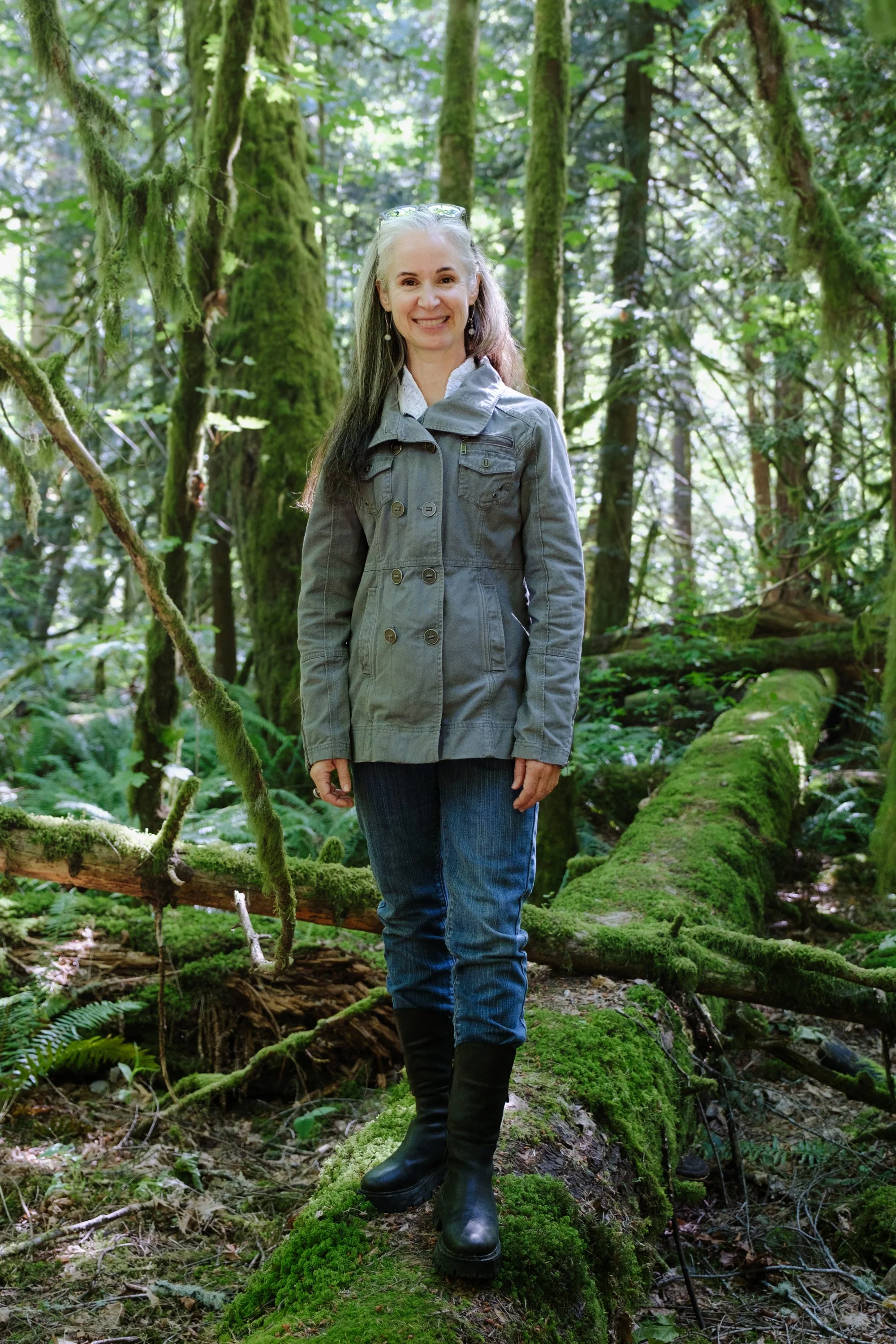 A smiling woman standing on a moss-covered log in a dense, green forest with tall trees and lush foliage.