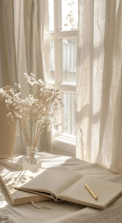Une fenêtre lumineuse avec des rideaux en lin, une vase avec des fleurs blanches, un carnet ouvert avec un stylo doré.
