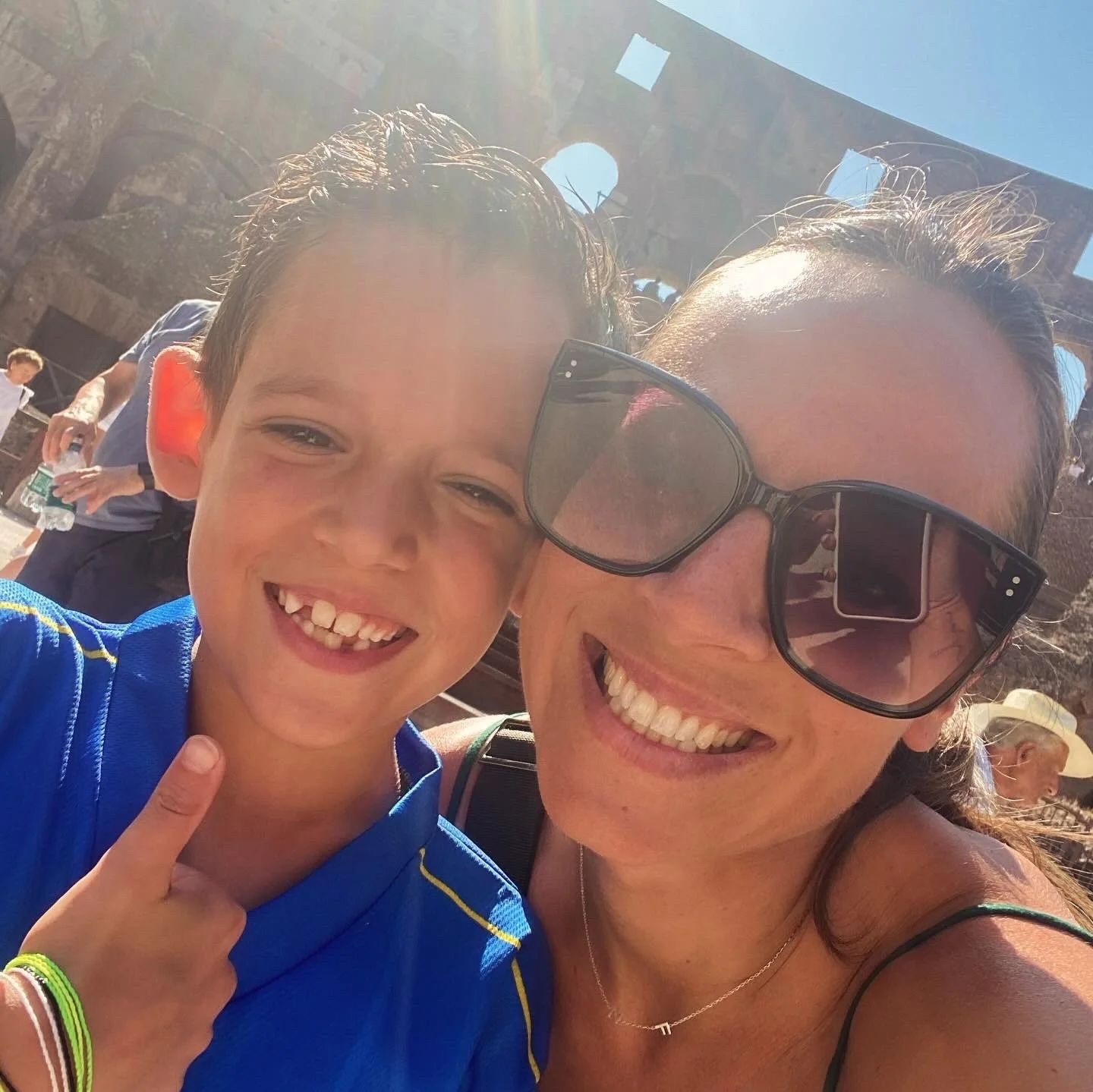 Une femme et un garçon souriants prennent un selfie à l'extérieur avec le monument du Colisée en toile de fond, ensoleillé avec un ciel bleu.