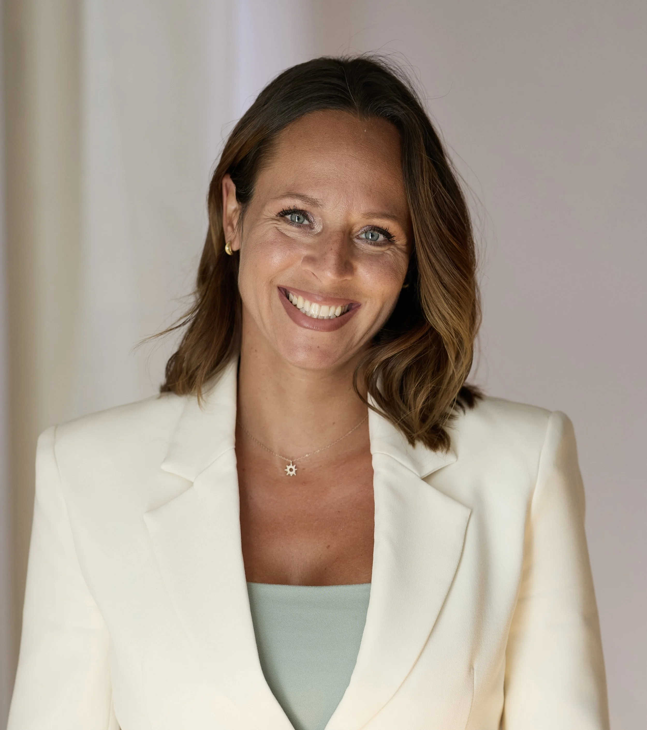 Une femme souriante avec des cheveux bruns, portant une veste blanche et un collier, dans un intérieur lumineux.