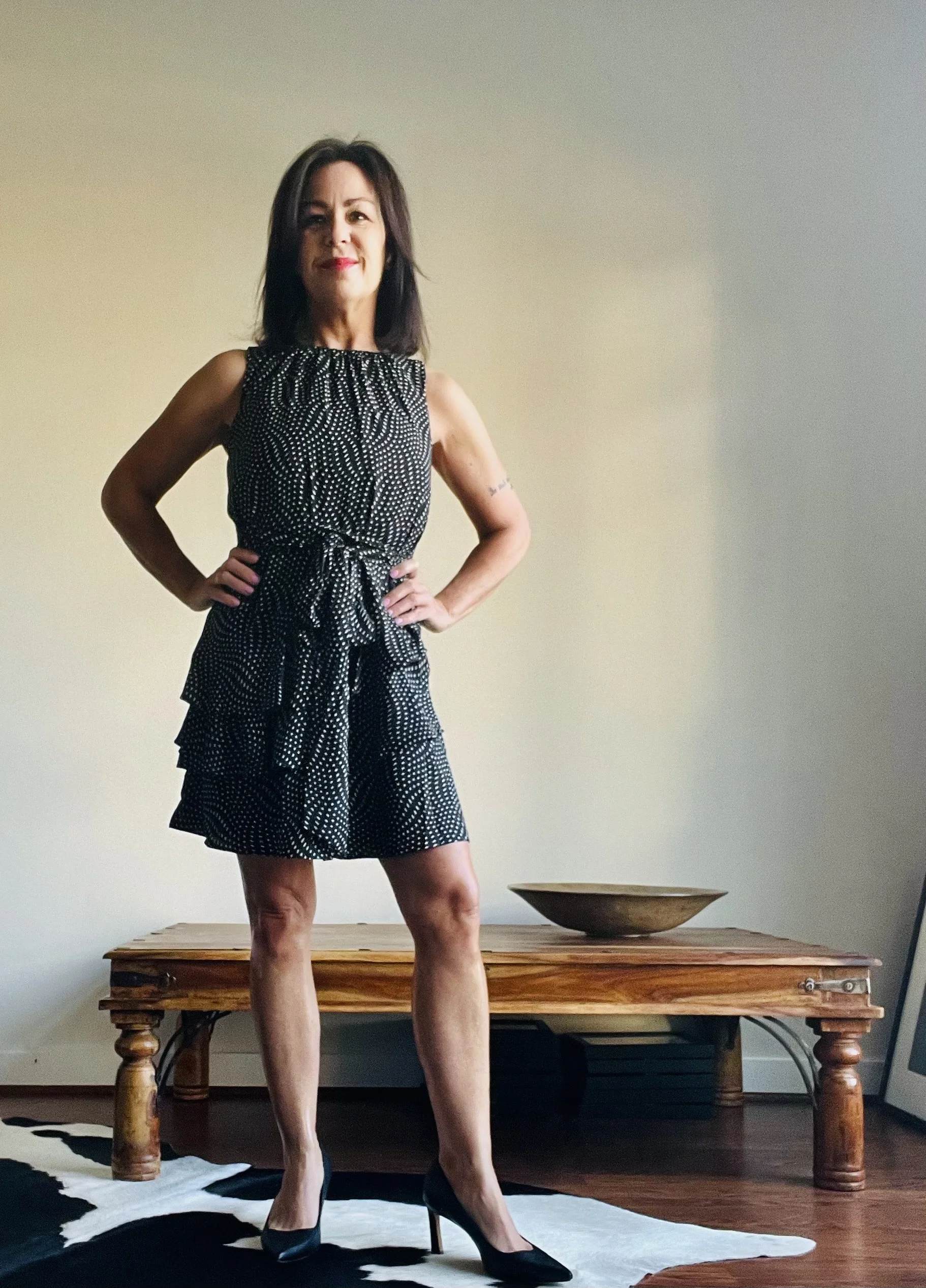 A woman with dark shoulder-length hair standing in a room, posing confidently with her hands on her hips. She is wearing a black dress with white polka dots, a waist tie, and black high heels. There is a wooden table and a bowl in the background, and she is standing on a black and white patterned rug.