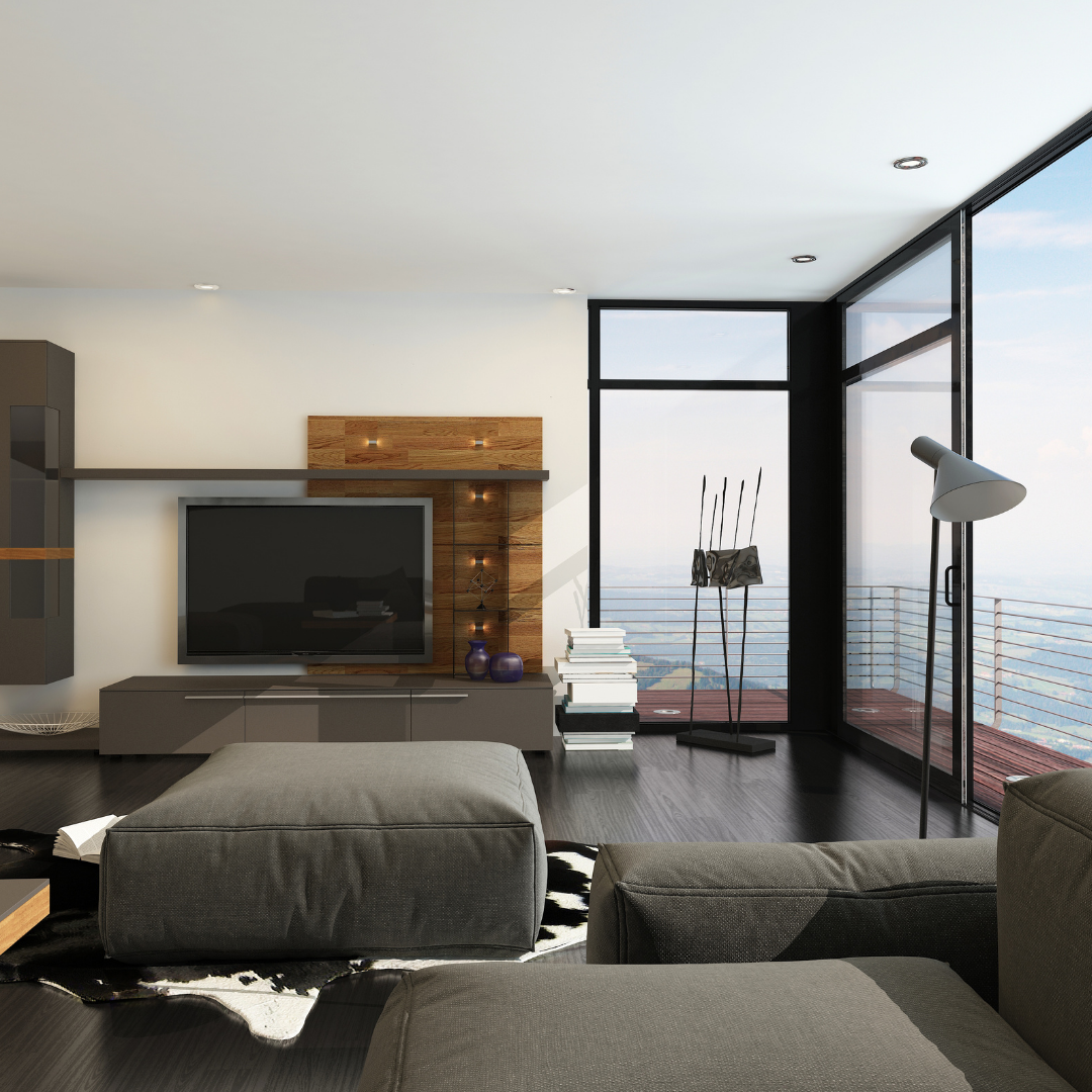 Modern living room with large glass windows, a TV on a wooden wall panel, a gray ottoman, a sofa, stack of books, and floor-to-ceiling balcony with a view of the city sky.