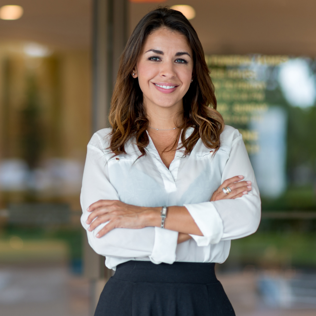 A woman with long brown hair is smiling with her arms crossed. She is wearing a white blouse and a dark skirt, standing indoors with a blurred background.