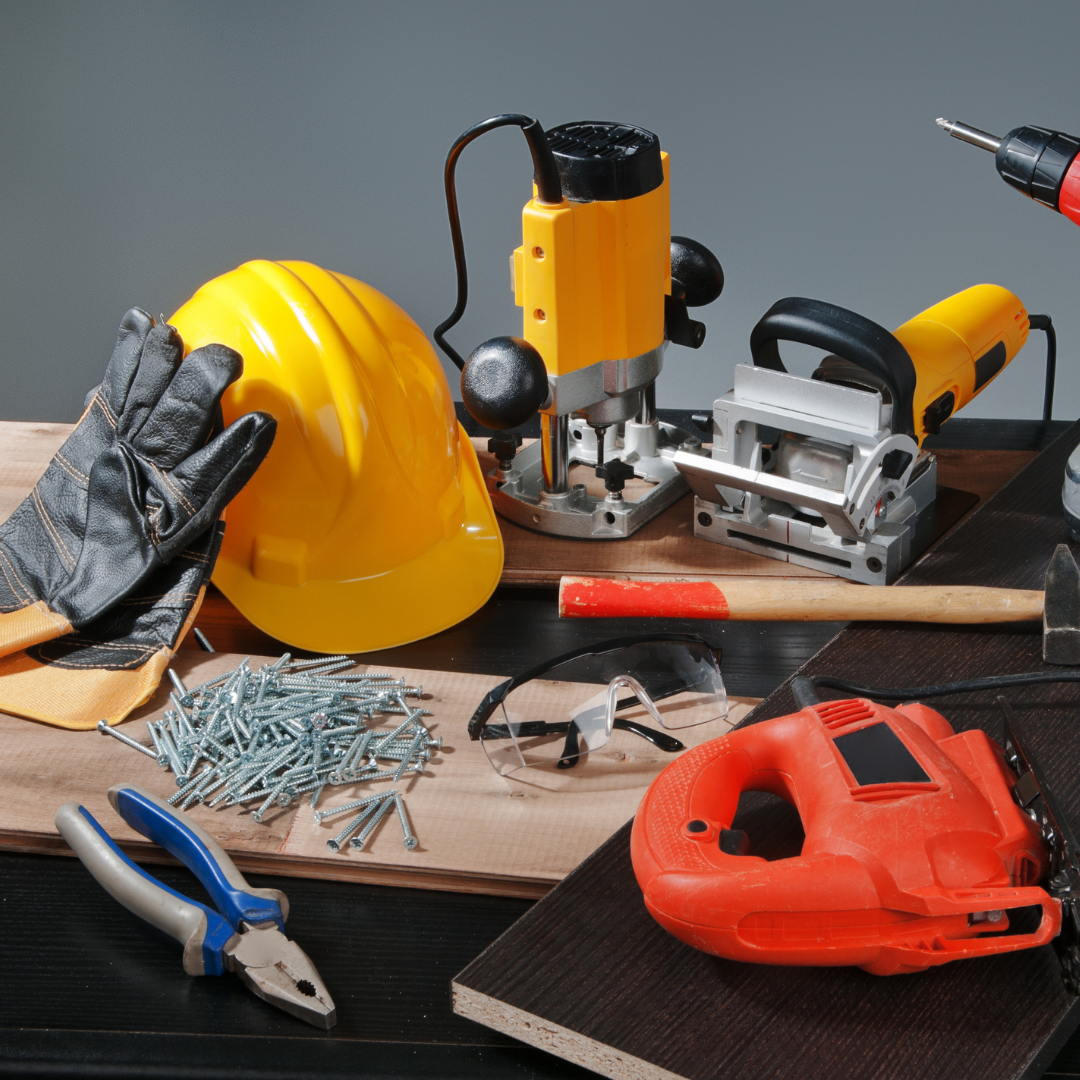 A collection of construction tools and safety equipment including a yellow hard hat, black gloves, a pair of safety glasses, a hammer, a pile of screws, a utility knife, a circular saw, a drill, and a cordless power saw, arranged on a wooden and black surface.