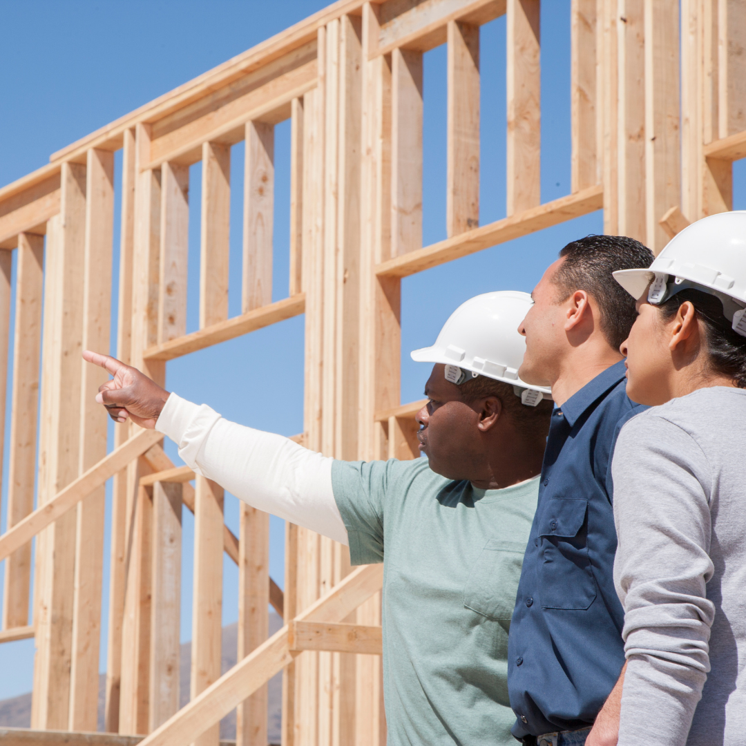 Three construction workers wearing white safety helmets review a wooden framing structure at a building site under a clear blue sky.