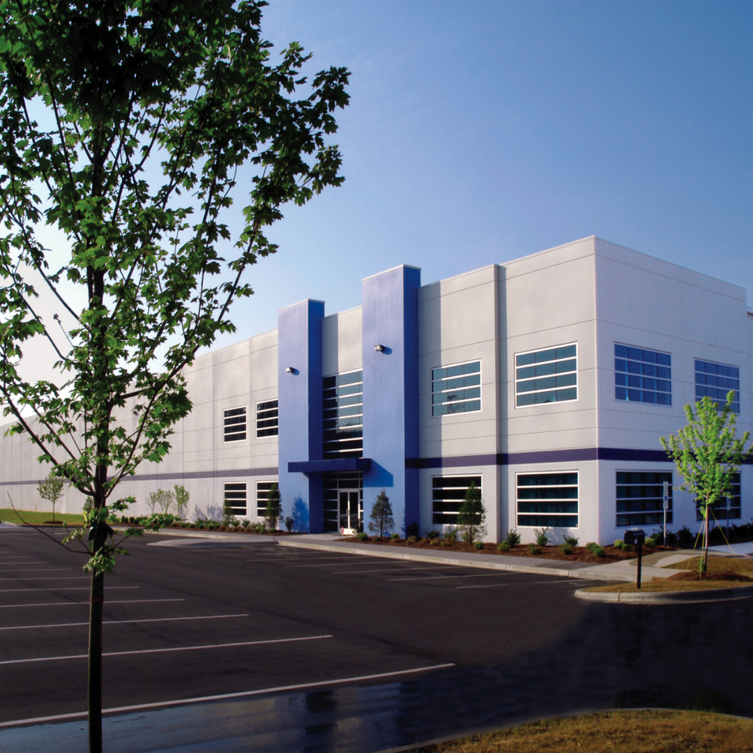 Modern commercial or industrial building with a parking lot in front, two trees, and a clear blue sky.