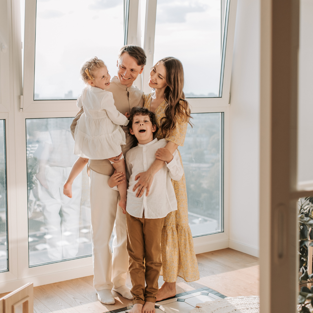 Happy family of four standing in front of large windows, smiling and embracing indoors on a sunny day.