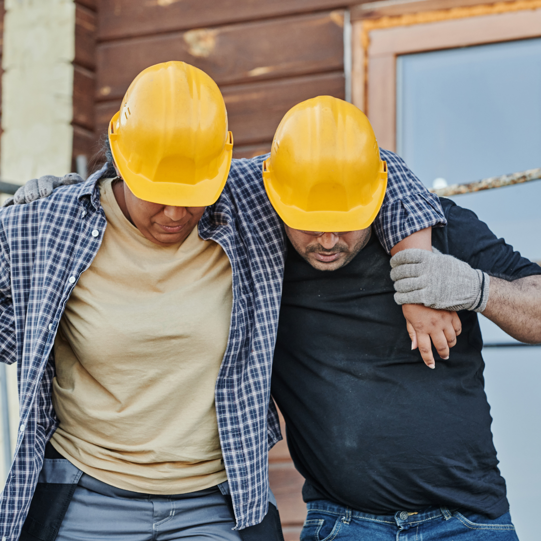 Two construction workers wearing yellow safety helmets helping each other walk, one with a checked shirt and the other with a black shirt, outdoors in front of a wooden building.