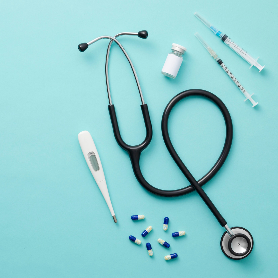 Collection of medical supplies including a stethoscope, digital thermometer, pills, vaccine vial, and syringes on a light blue background.
