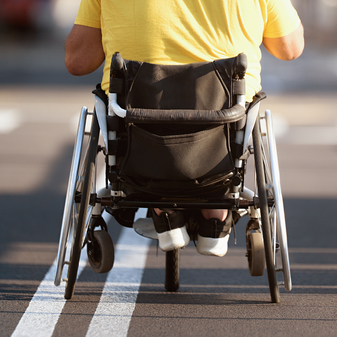 Back view of a person in a wheelchair wearing a yellow shirt, on a marked path outdoors.