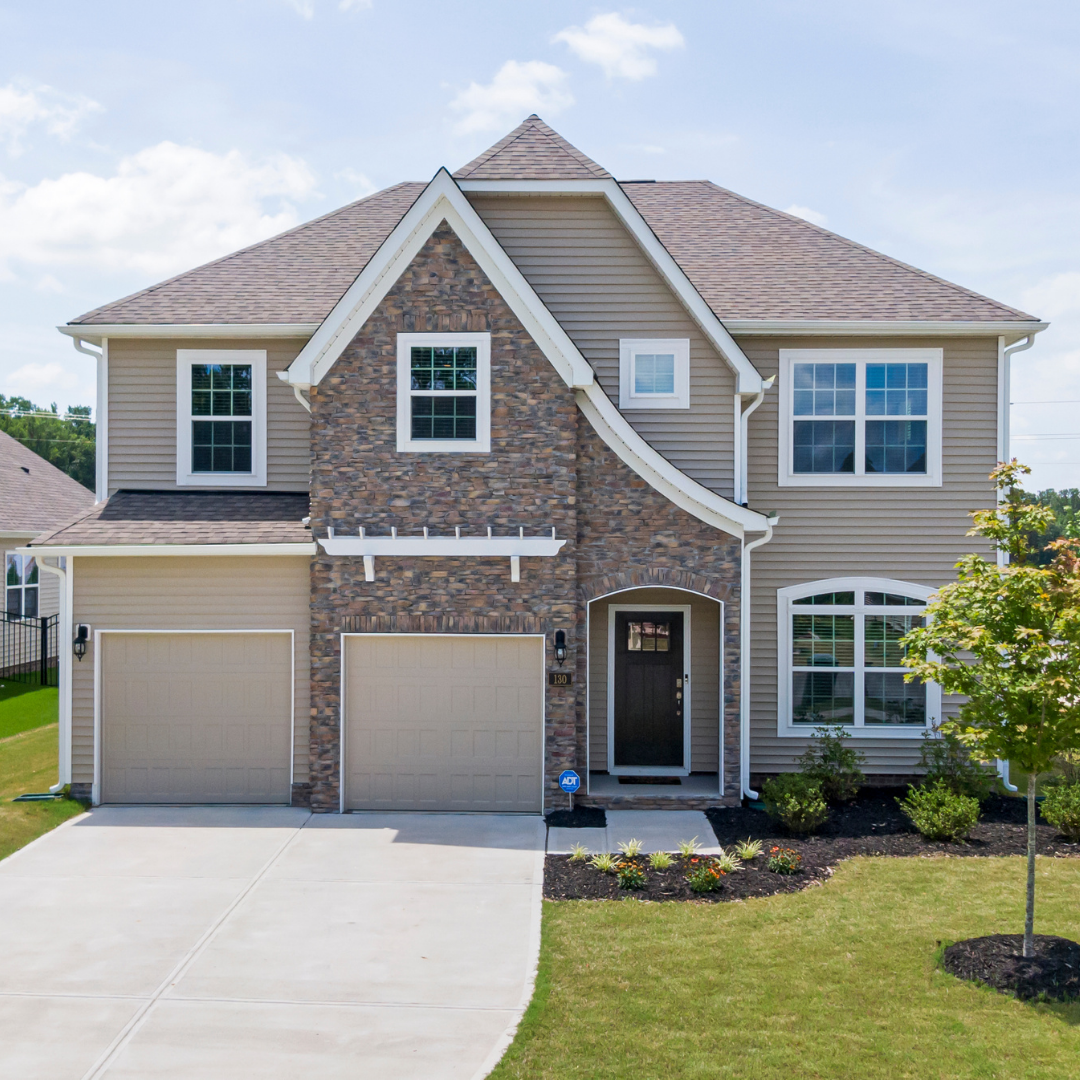 Front view of a two-story house with beige siding, a stone facade, and a dark front door, surrounded by a lawn and landscaping, with a driveway leading to a two-car garage.