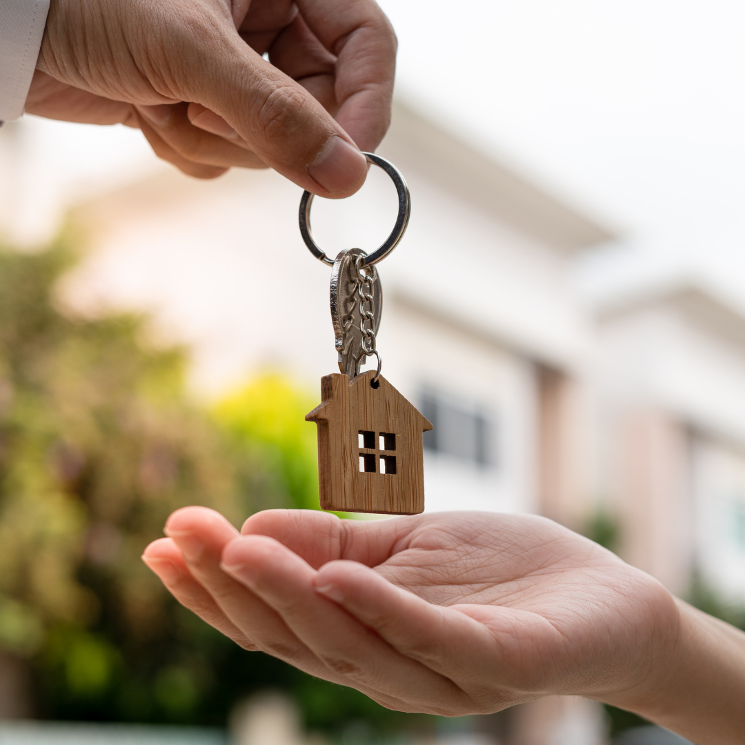 A hand holding a house-shaped keychain, extending it over another hand. The background shows a residential neighborhood with lush greenery.