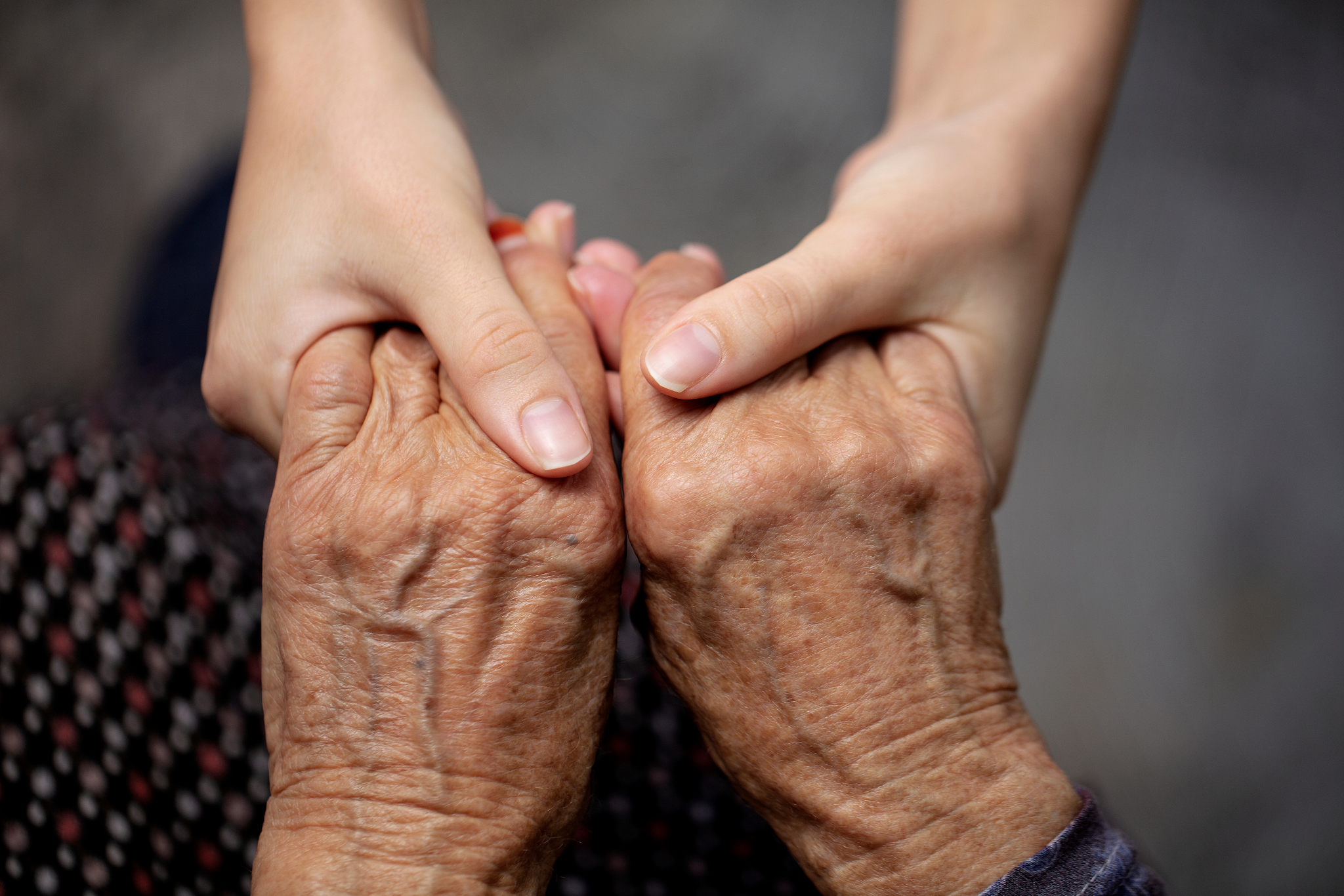 Two hands, one young and one elderly, holding and clasping each other's fingers in a gesture of care and support.