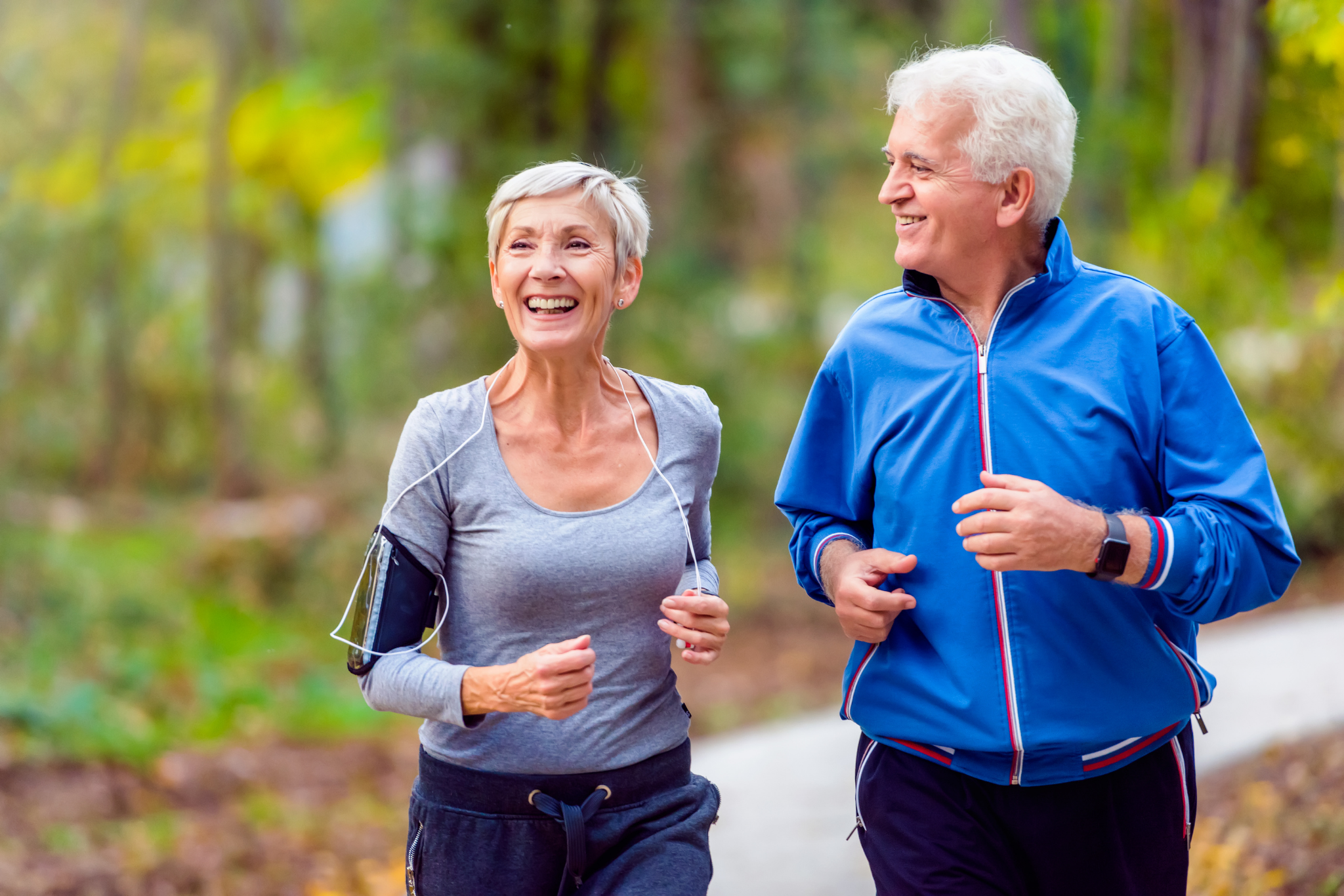 An elderly couple jogging in a forested park, smiling and enjoying their workout.