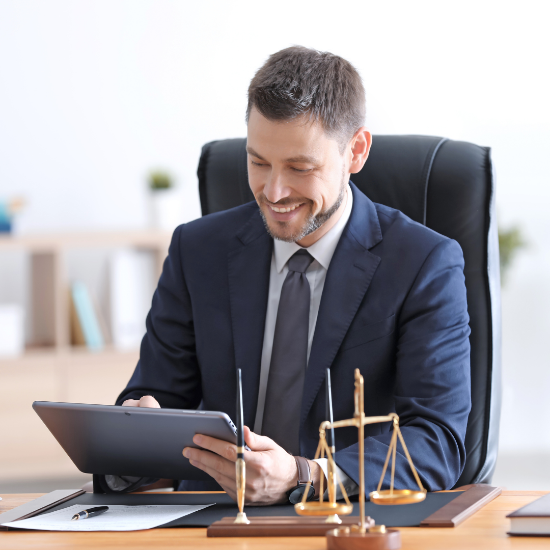 A smiling man in a navy suit and tie sitting at his desk, looking at a tablet, with a decorative brass scale on the desk.