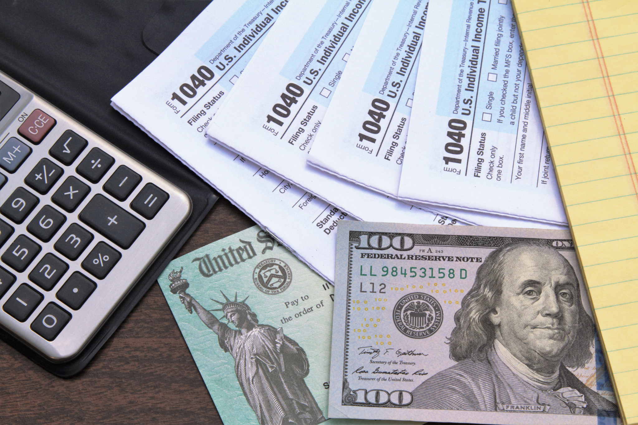 The image shows a calculator, three federal income tax forms 1040, a hundred dollar bill, a twenty dollar bill, and a yellow notepad on a dark wooden surface.