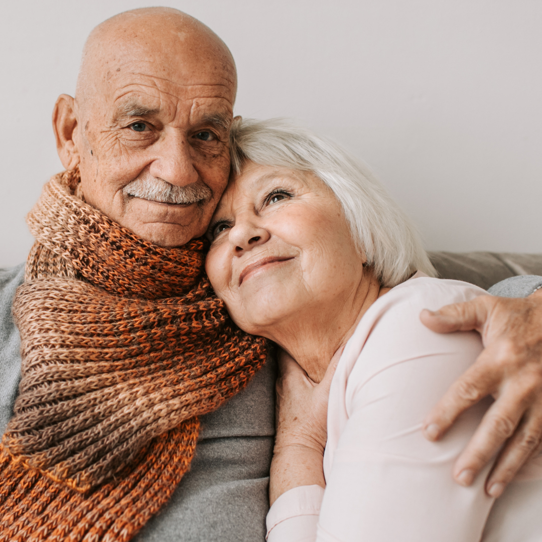 An elderly couple sits close together, smiling and hugging each other affectionately.
