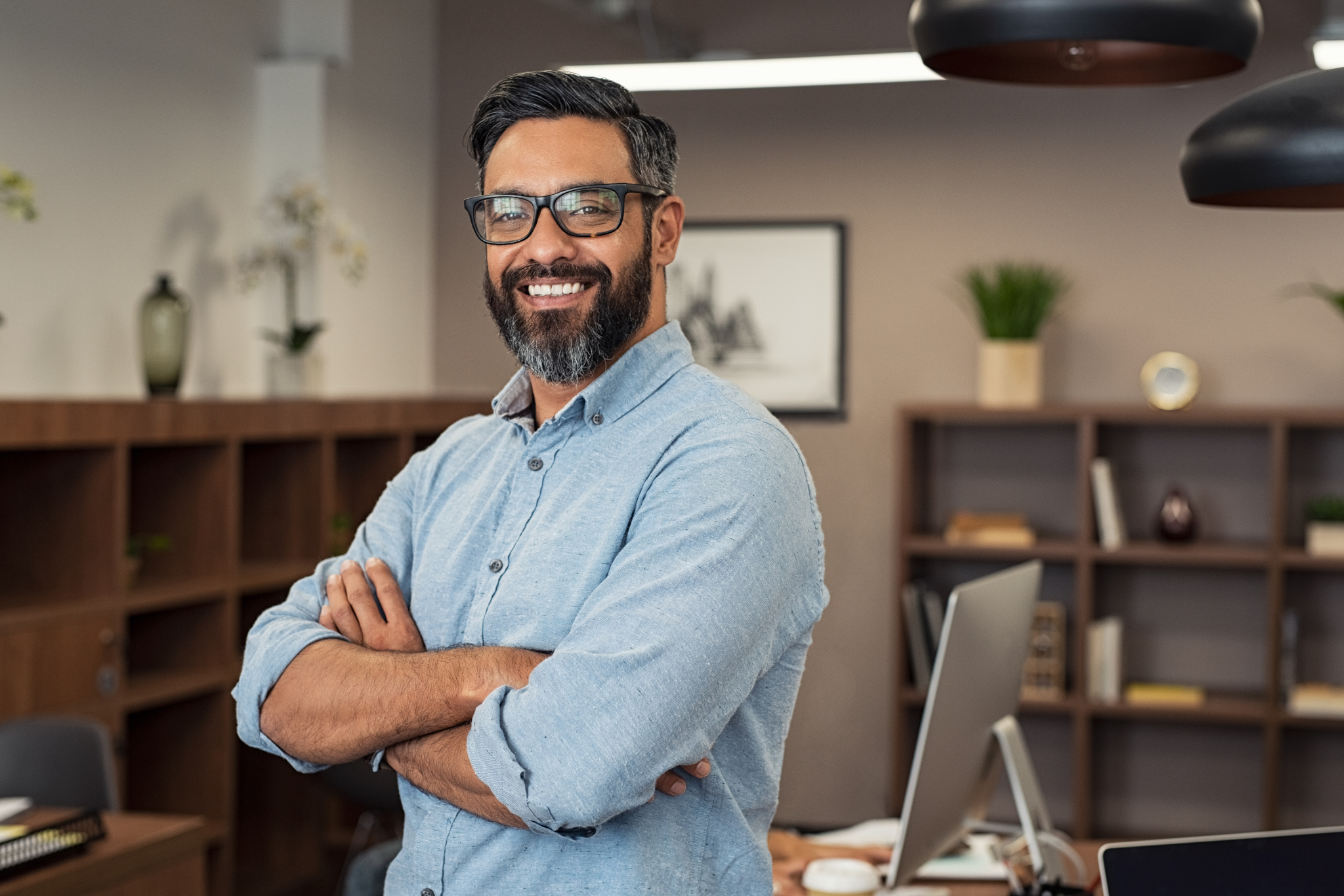 A confident man with glasses, beard, and dark hair, standing with arms crossed in an office, smiling at the camera.