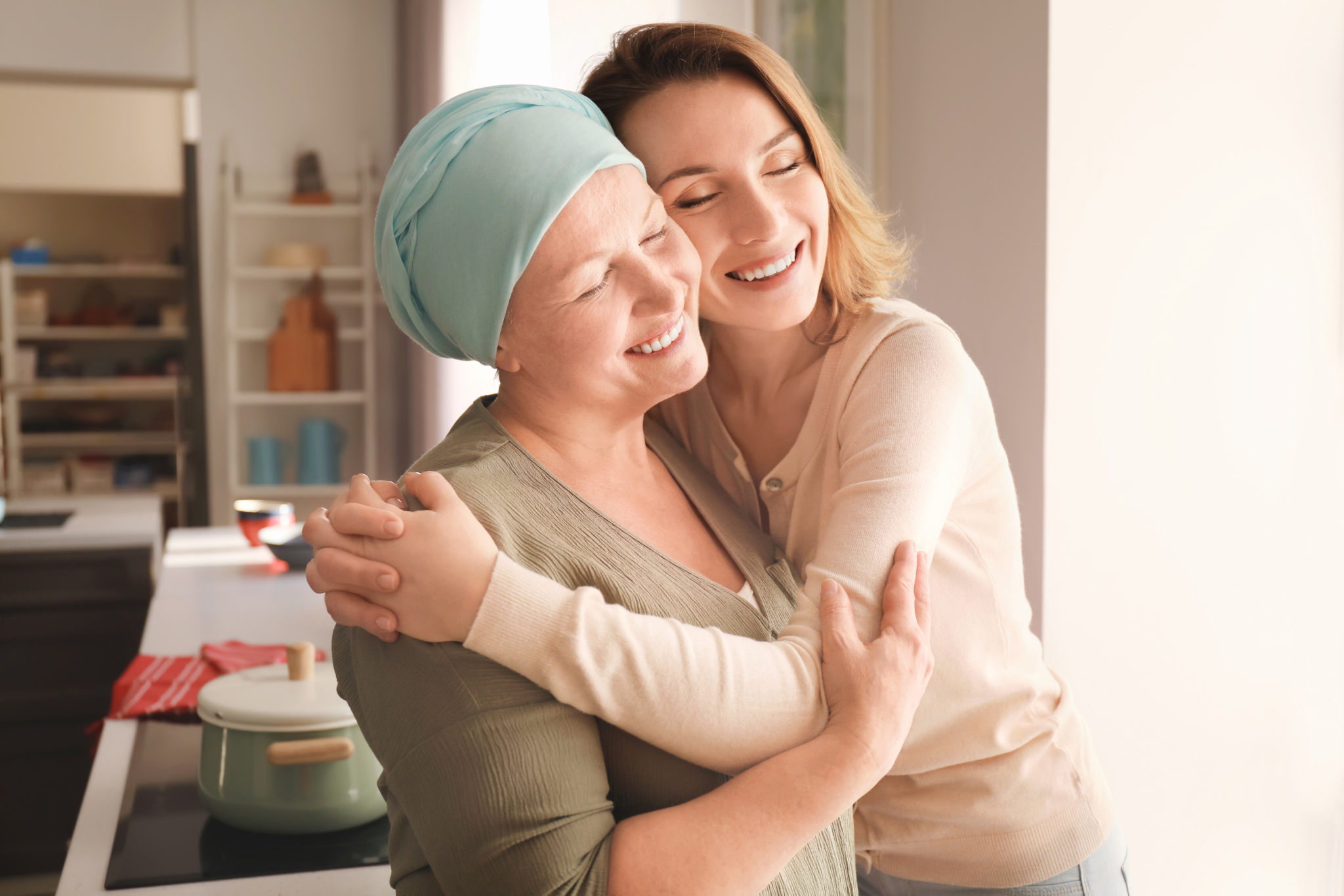 A young woman hugging an older woman with a light blue headscarf, both smiling happily in a kitchen.