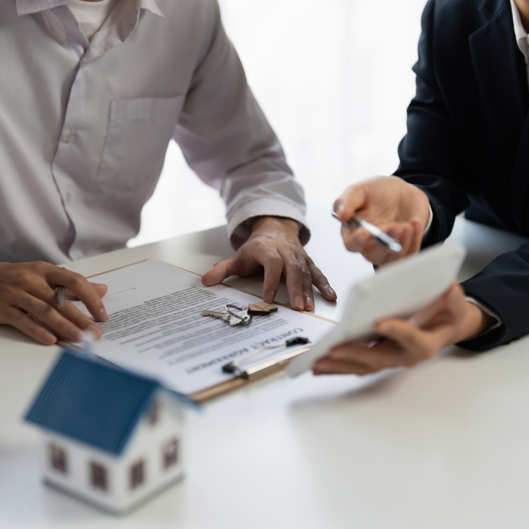 Two people reviewing a contract document with a small house model on the table, indicating a real estate transaction or property discussion.
