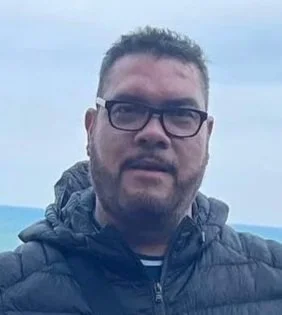 A man wearing glasses and a blue jacket, outdoors with cloudy sky in the background.