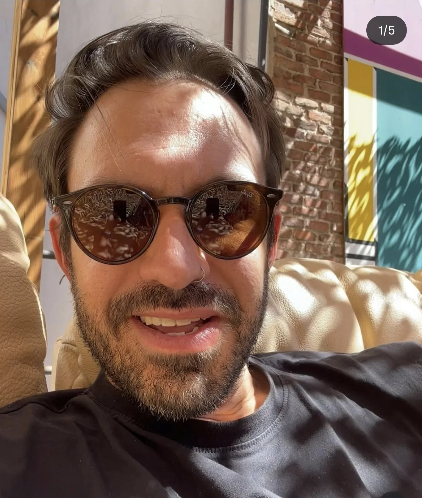 A man with dark hair, a beard, and sunglasses, sitting on a tan leather couch indoors, with a brick wall and colorful abstract artwork in the background.