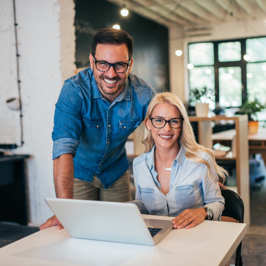 A man and woman wearing glasses smiling at a laptop in a modern office