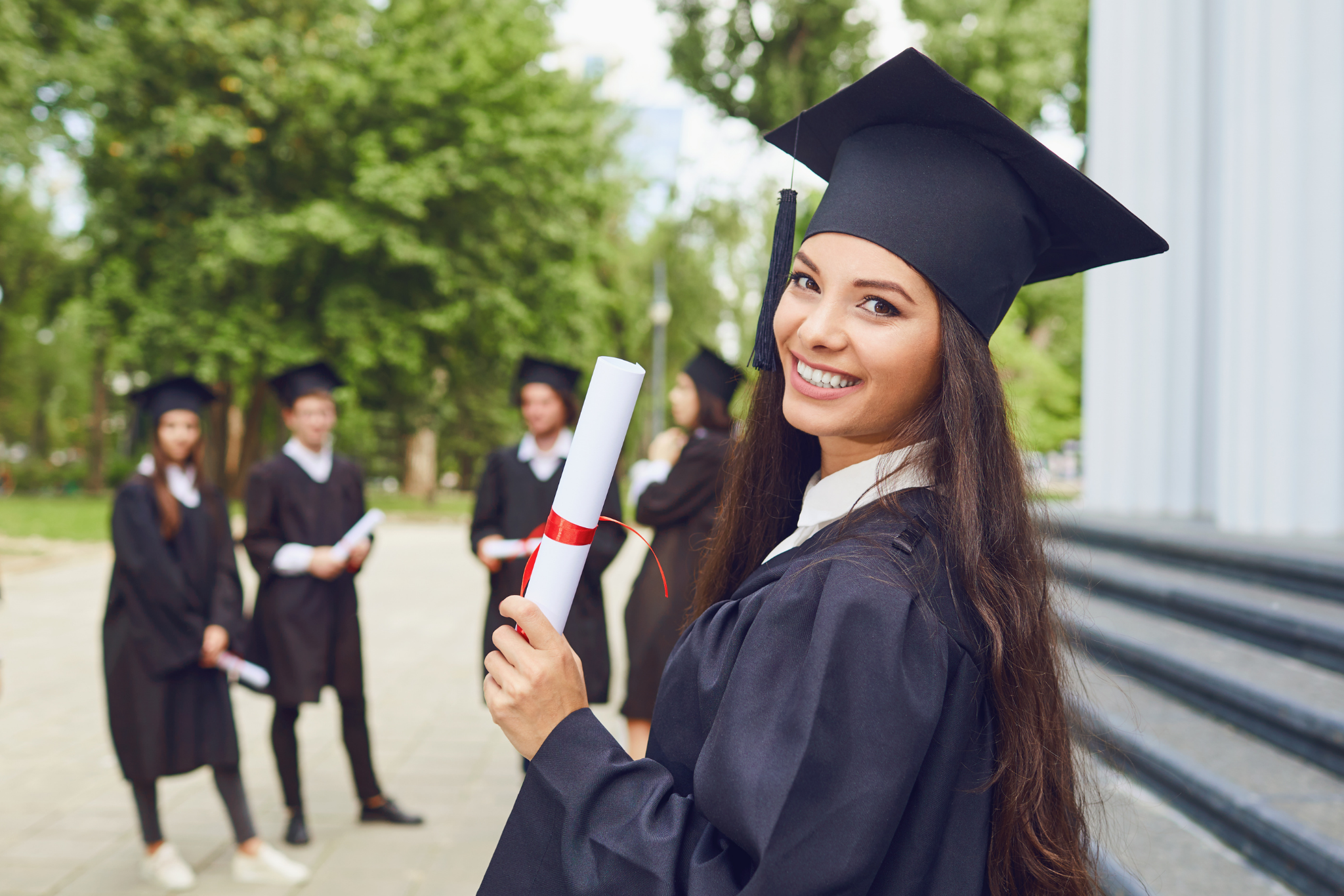 Young woman in graduation cap and gown smiling and holding a diploma, standing outdoors with other graduates in the background.