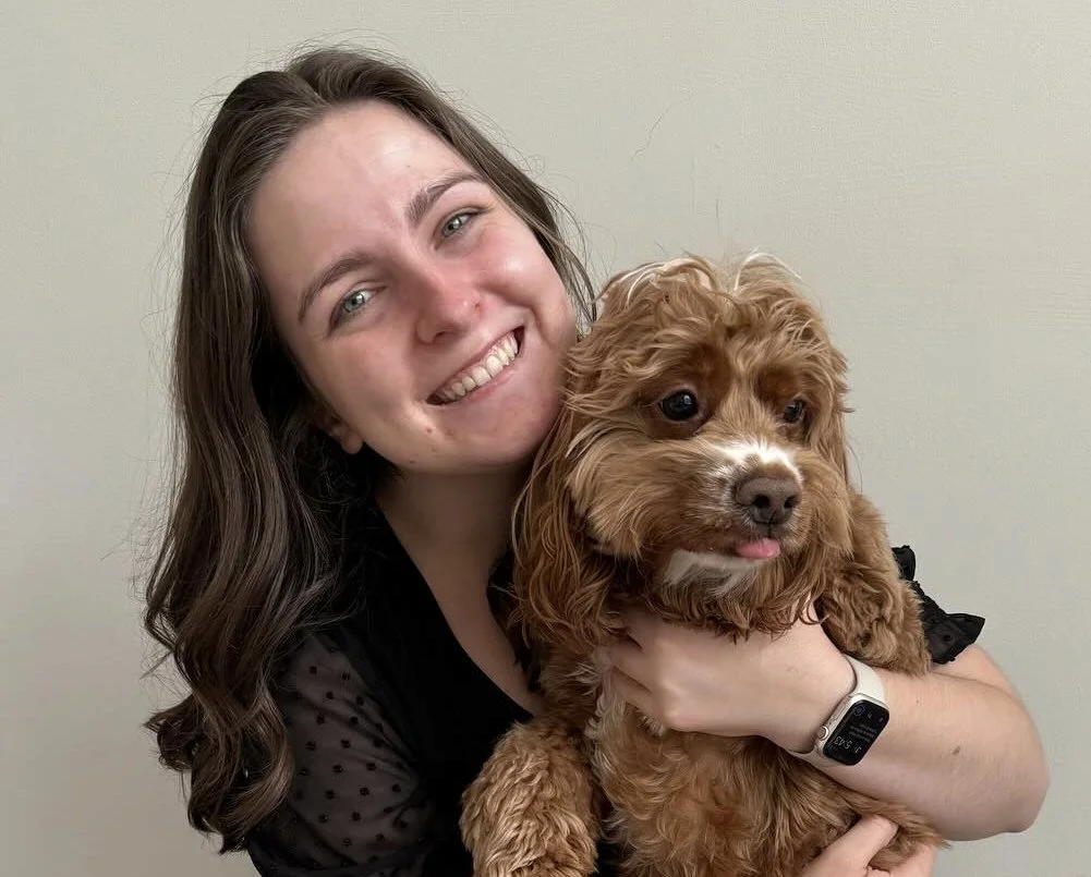 A woman with long brown hair smiling and holding a small brown curly-haired dog in her arms.