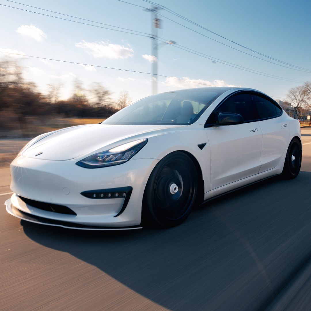 A white Tesla Model 3 electric car driving on a highway during the daytime with a clear sky and some clouds.
