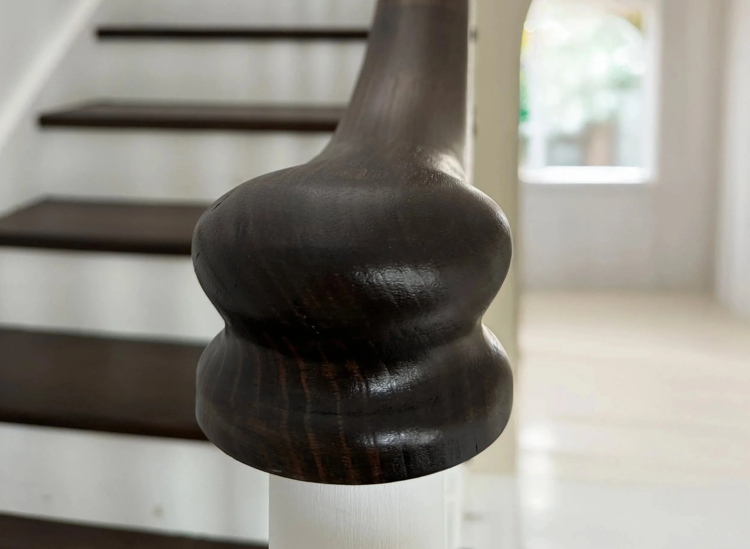 Close-up of a dark wooden staircase banister with a rounded top, inside a bright room with a staircase and a window in the background. Staircase Polishing Sydney