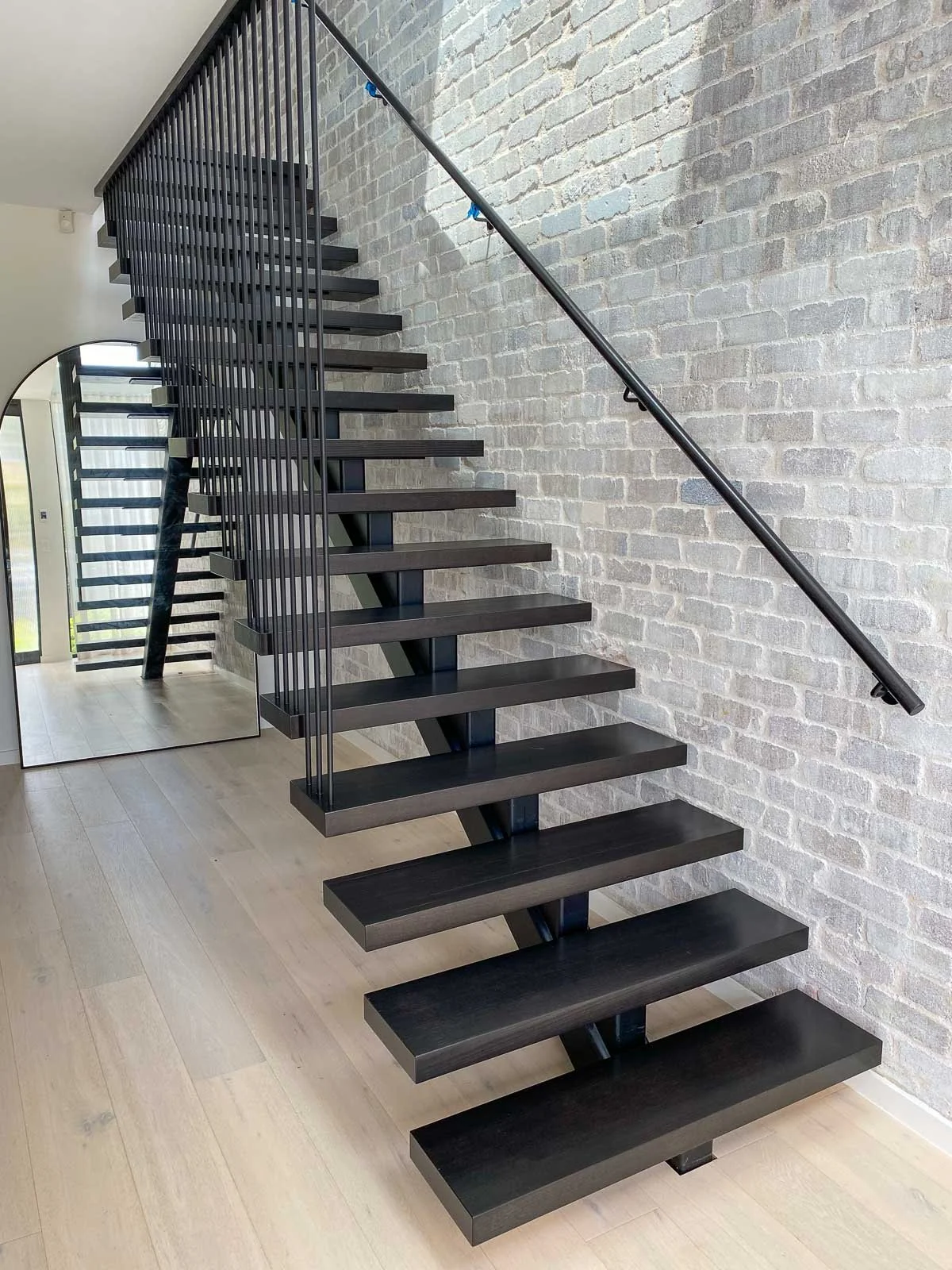 Modern black staircase with open steps and a simple black handrail located against a white brick wall inside a contemporary home. Staircase Polishing Sydney