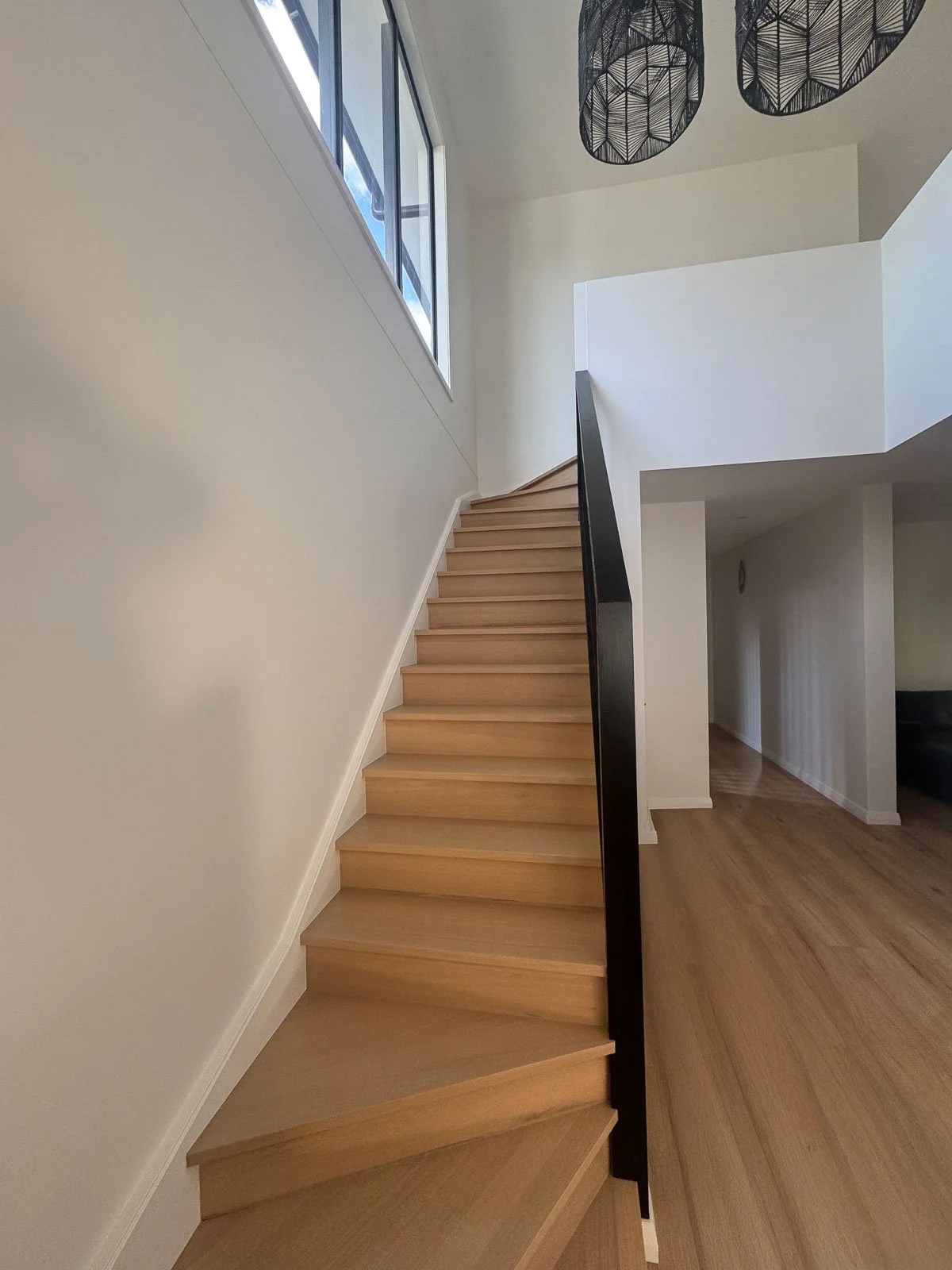 Wooden staircase with black railing leading to upper floor, white walls, a large window, and modern black pendant lights hanging from the ceiling.