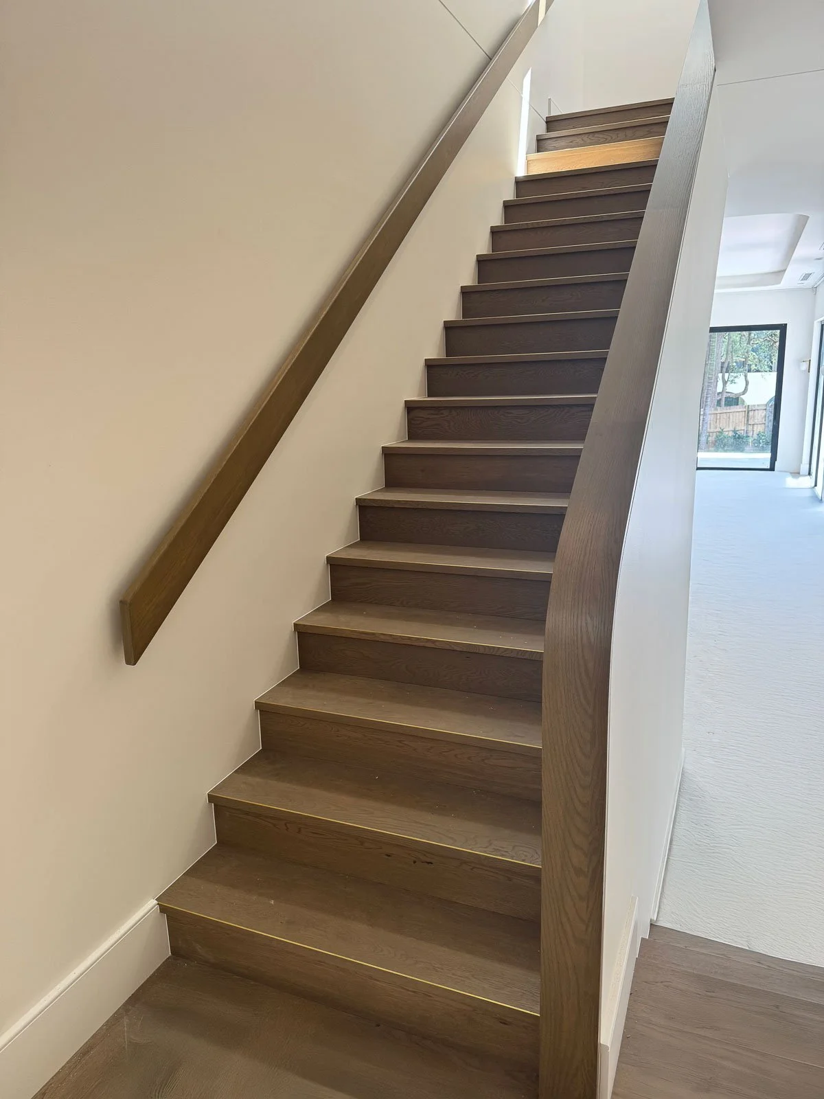Wooden staircase with a handrail on the left and a curved wall on the right, leading upward in a well-lit interior space.