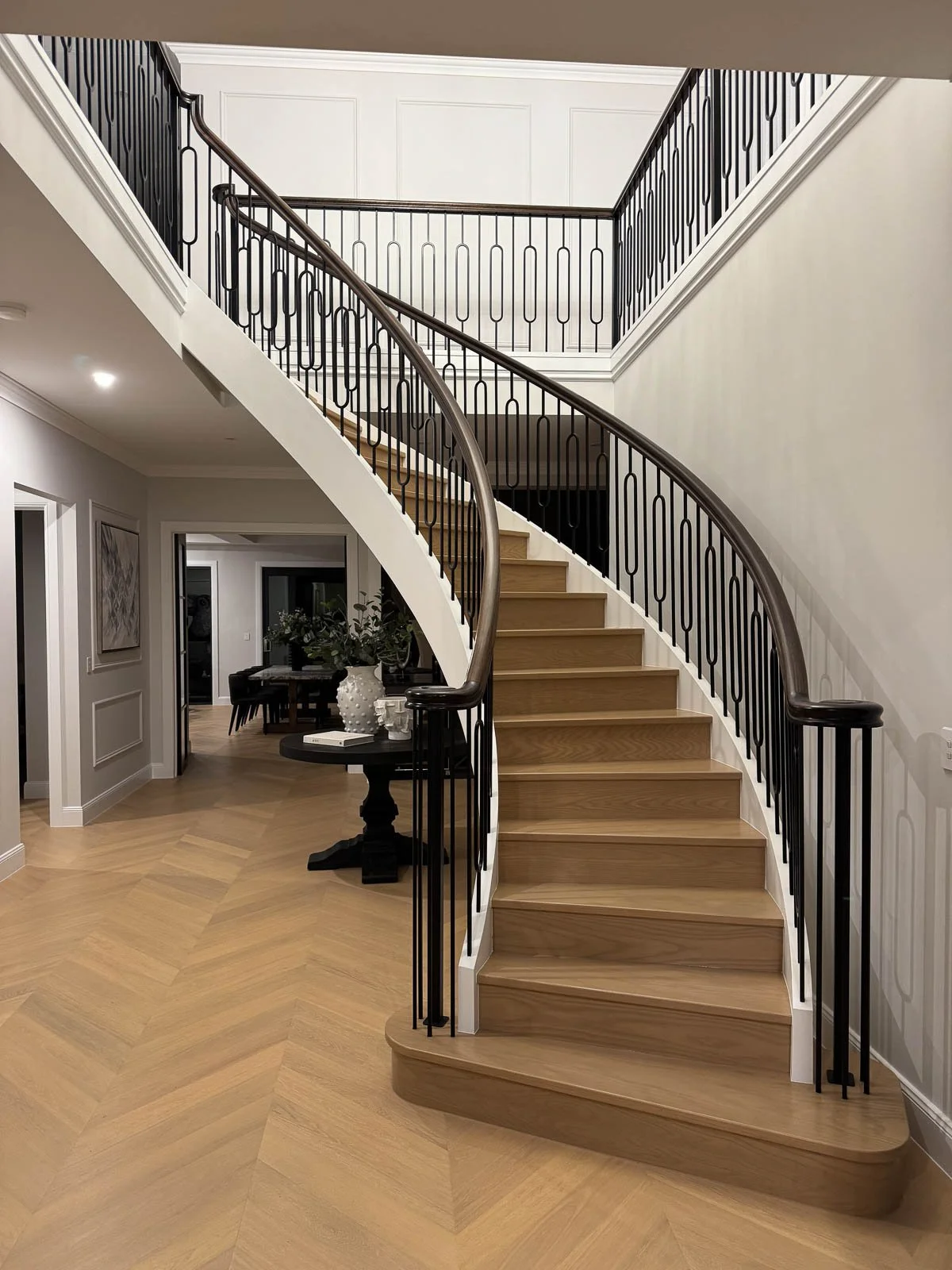 A curved staircase with wooden steps and black metal railings inside a modern home, leading to a second-floor balcony. Staircase Polishing Sydney