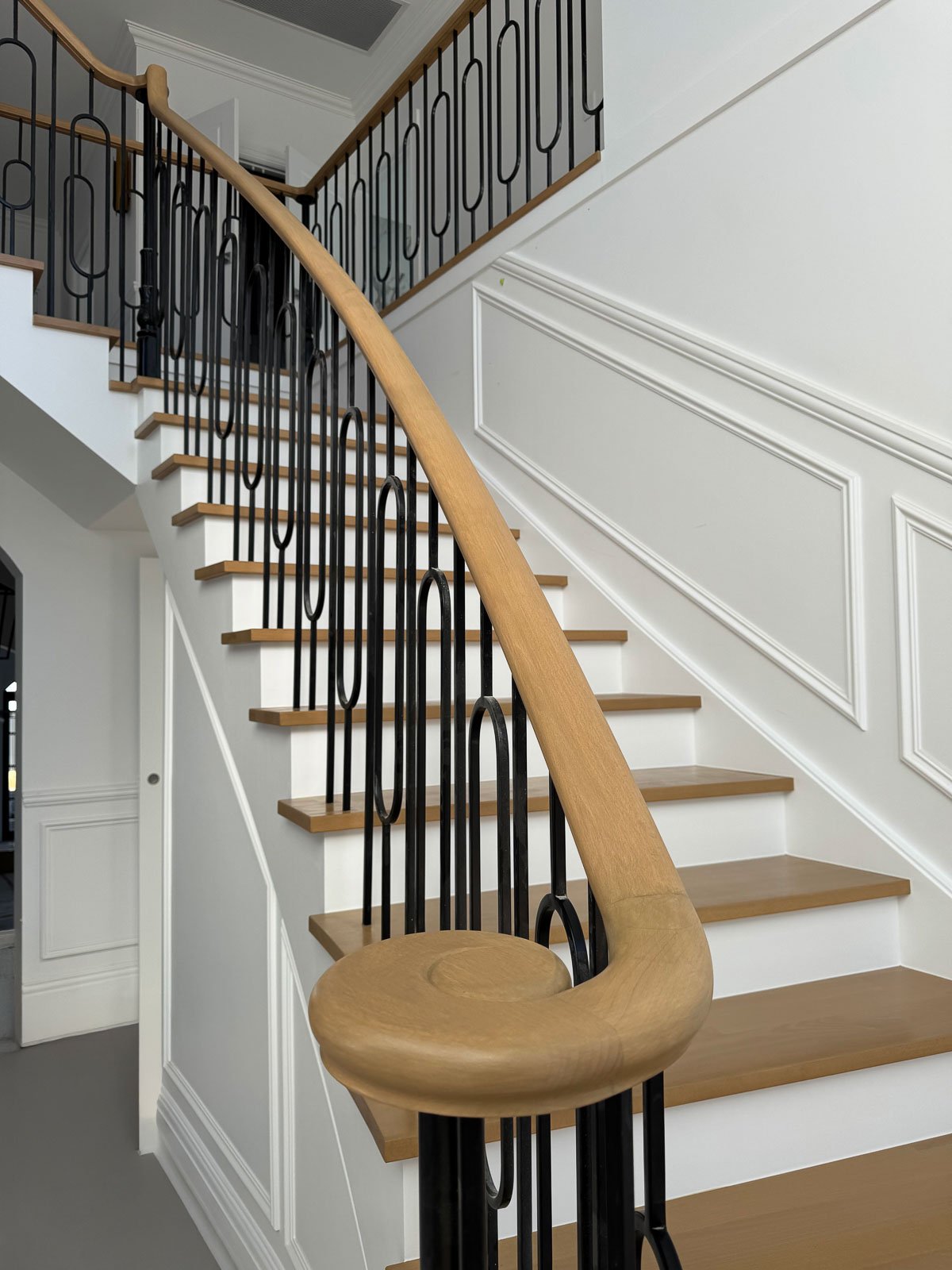 Interior staircase with wooden handrail, black metal balusters, white wainscoting, and light gray carpeted steps.