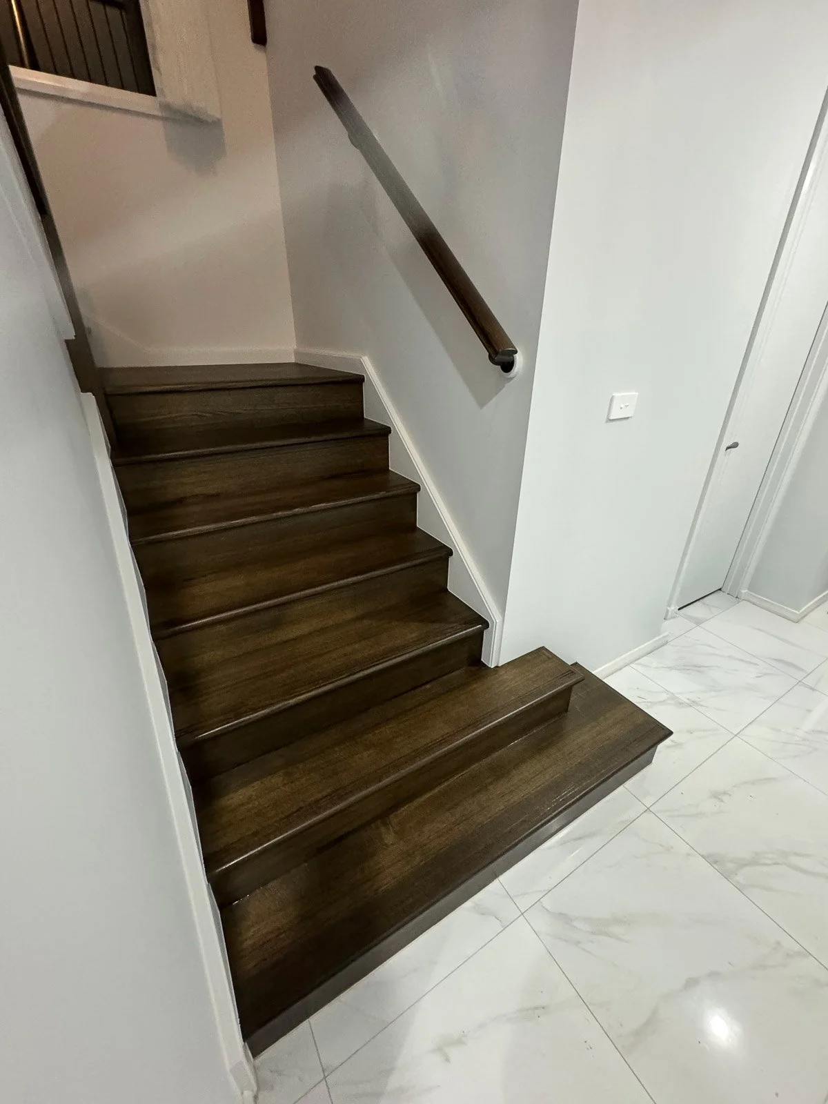 Wooden staircase with a dark finish leading upstairs, with a handrail on the right side, next to white walls and a light-colored tile floor.