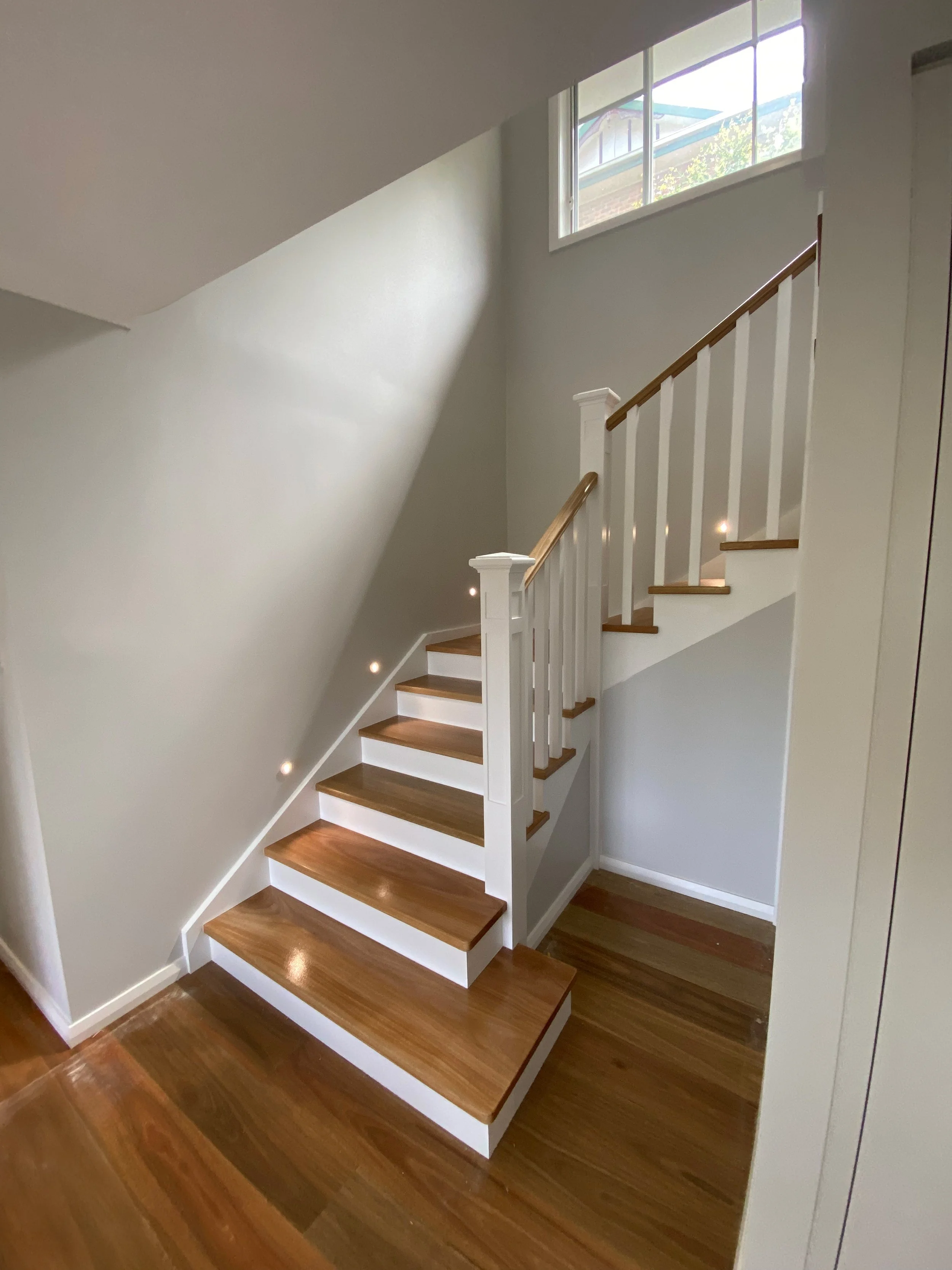 Interior staircase with wooden steps, white risers, white railing with wooden handrail, next to a wall with smart lights, and a large window bringing in natural light. Staircase Polishing Sydney