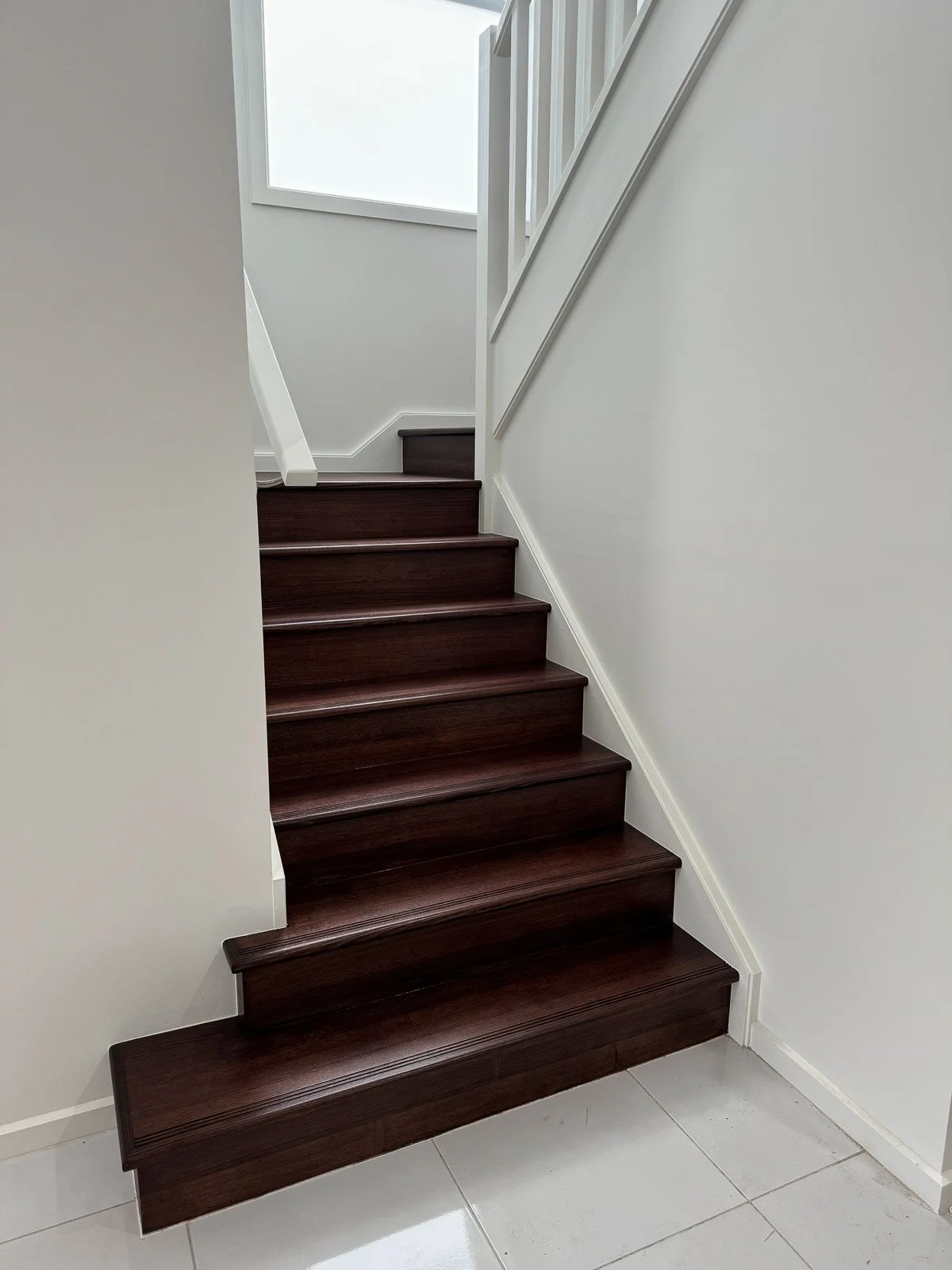 Interior view of a staircase with dark wood steps and white walls, leading upwards with a window at the top letting in natural light. Staircase Polishing Sydney