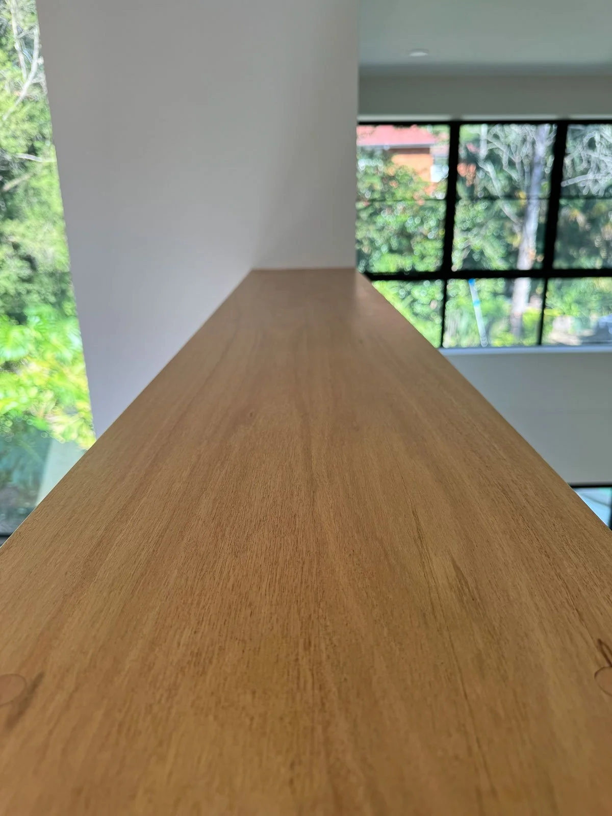 Close-up of a wooden railing or ledge inside a house, with large windows showing trees and greenery outside. Staircase Polishing Sydney